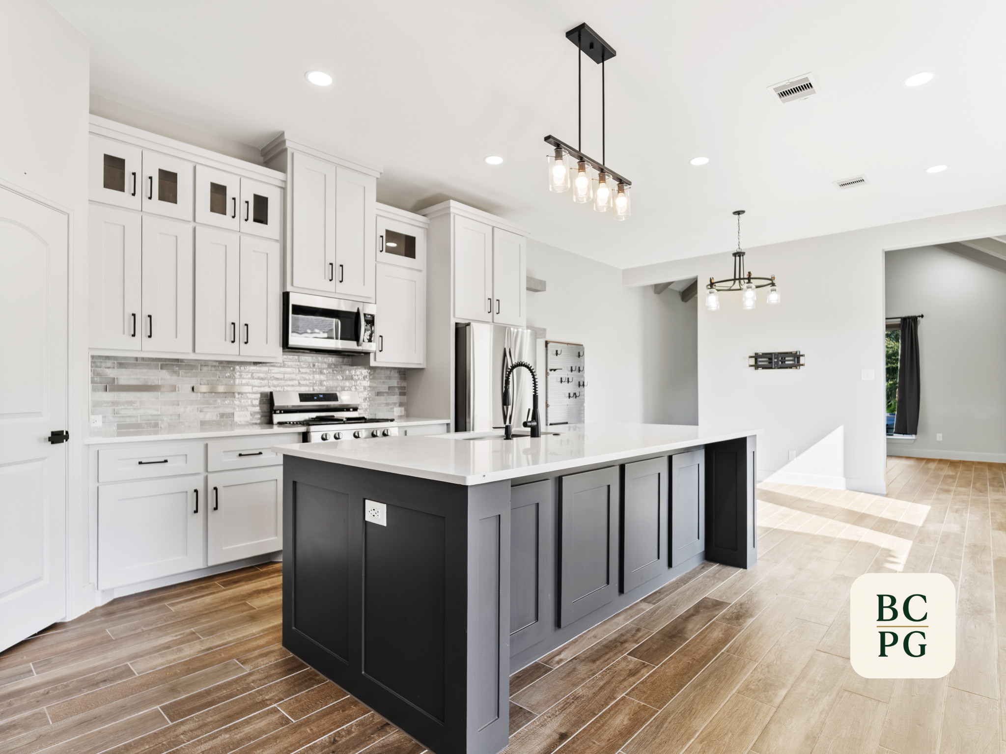 A modern kitchen with white cabinets, an island painted dark blue, and a light-colored tile floor. There are two hanging light fixtures with exposed bulbs, and stainless steel appliances including a microwave and a refrigerator. The space is bright w