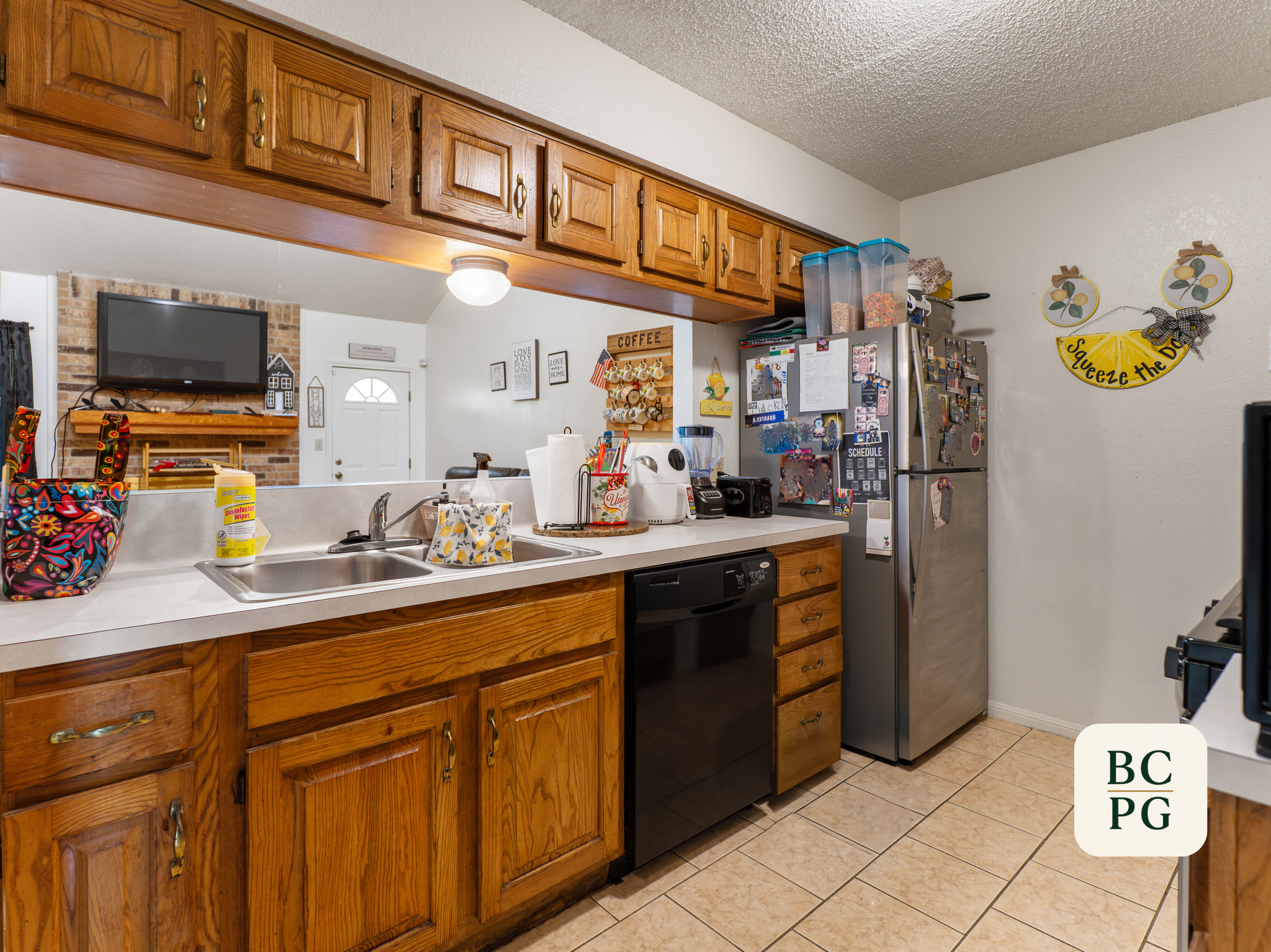 Kitchen with wooden upper cabinets, a stainless steel refrigerator decorated with photos and notes, a countertop with various kitchen items, and wall decorations including a yellow sign that says "Squeeze the Day".