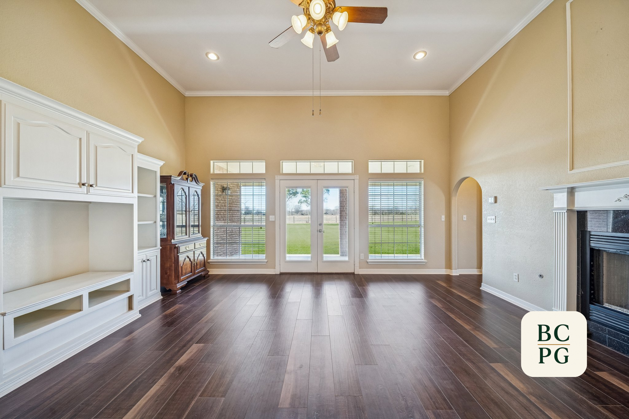 Living room with large windows, glass doors leading outside, beige walls, white crown molding, dark wood flooring, and a ceiling fan with lights.