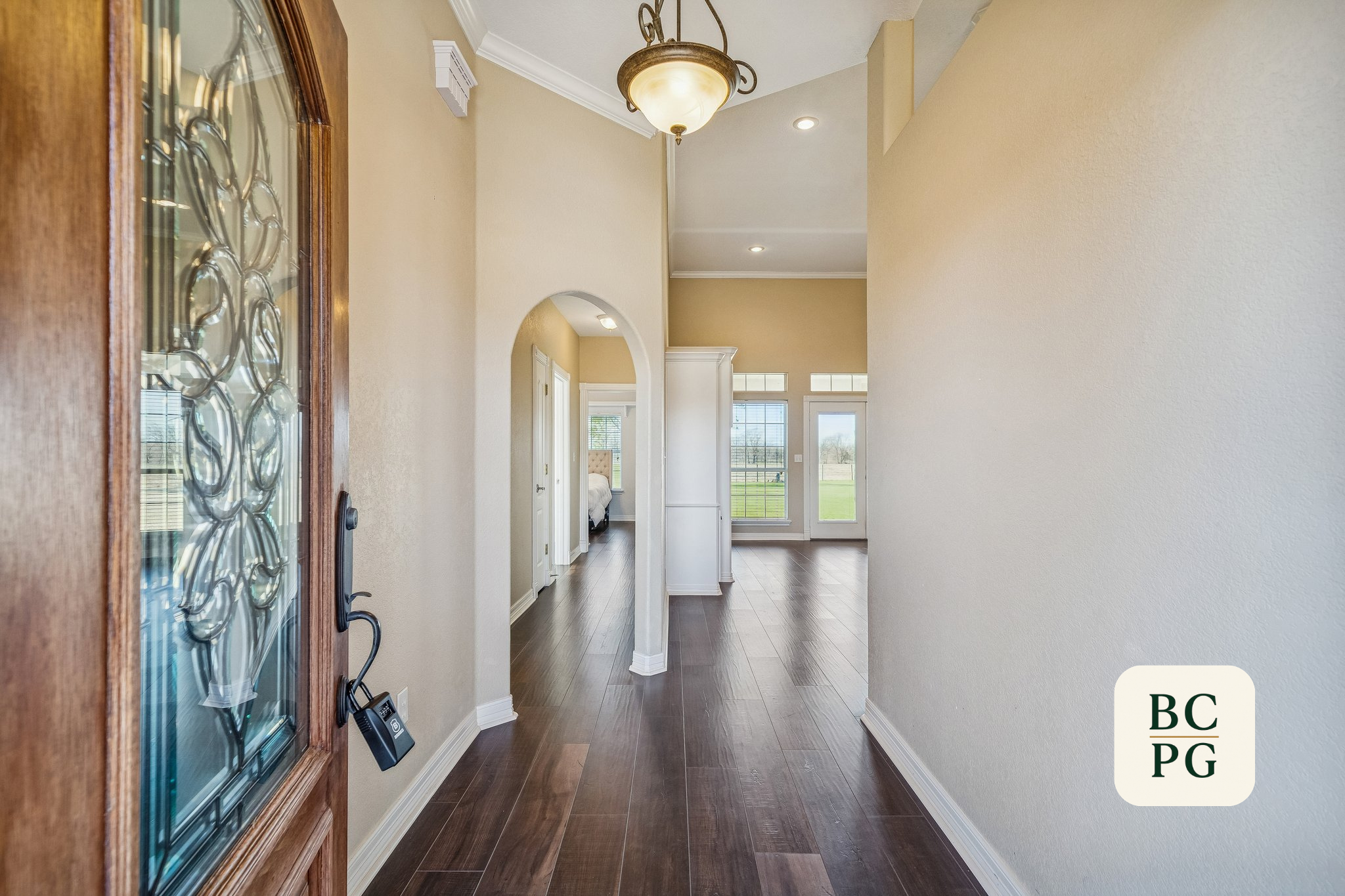 Interior hallway of a house with wooden floors, beige walls, and a white ceiling with crown molding. A chandelier with a frosted glass shade hangs from the ceiling. There are multiple windows at the end of the hallway showing a view of green fields o