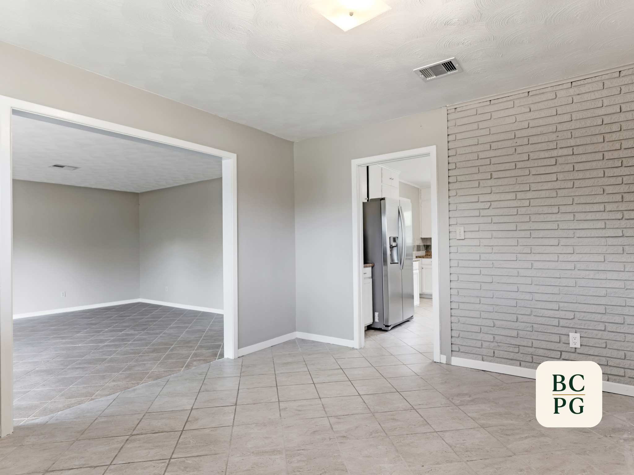 Living room and kitchen with tiled floors, white walls, and a brick accent wall. The living area has large open space with an open doorway leading to a kitchen with a stainless steel refrigerator.