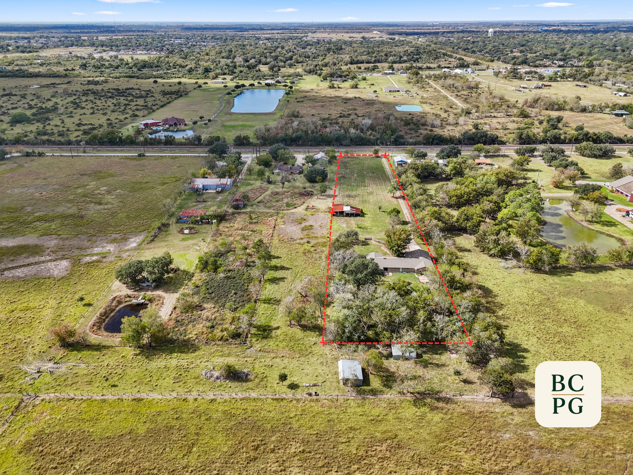 Aerial view of a rural landscape with houses, ponds, and open fields. There are areas marked with red dashed lines and labels indicating different sections.