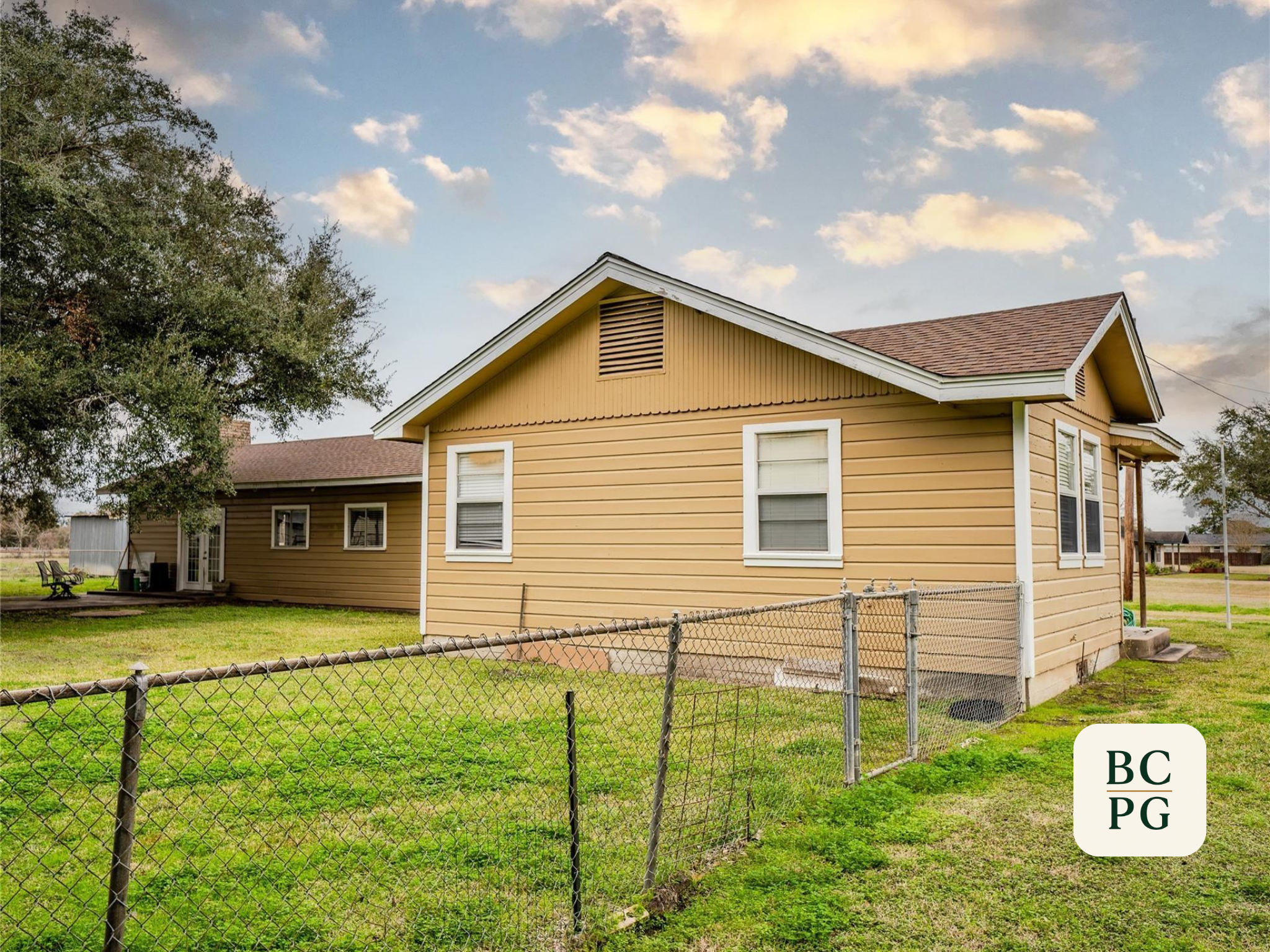 A yellow house with white trim and two windows, a backyard with a chain-link fence, a chairs, and a shed, under a partly cloudy sky.