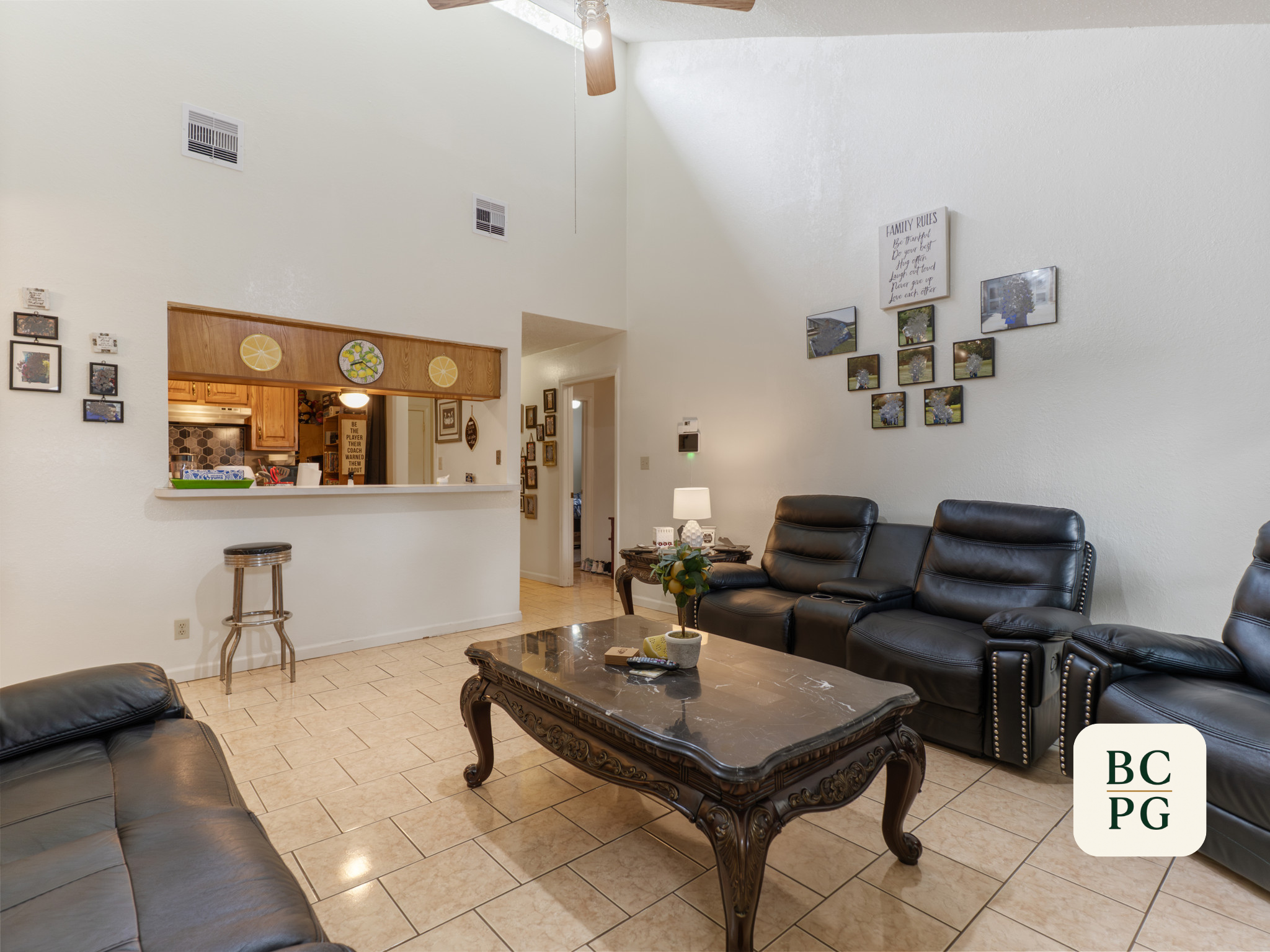 Living room with black leather sofas, a marble coffee table, a lamp, and family photos on the wall. An open kitchen with wooden cabinets is visible through a pass-through window.