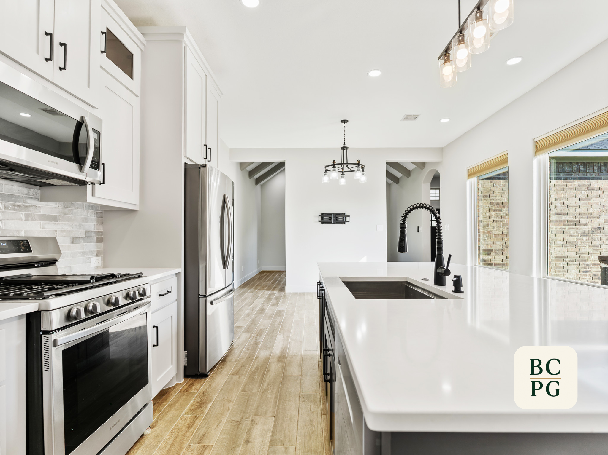 Modern white kitchen with stainless steel appliances, black fixtures, wooden flooring, and large windows with blinds.