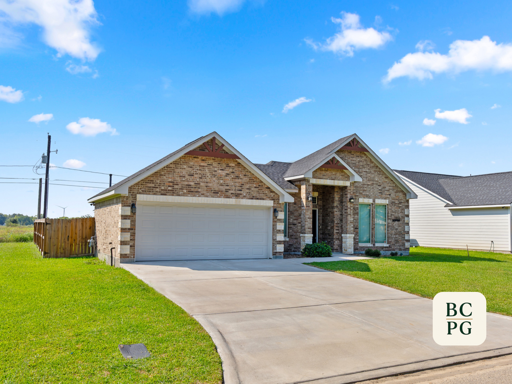 A modern brick house with a white garage door, a concrete driveway, a small neatly landscaped front yard, and a bright blue sky with a few clouds.
