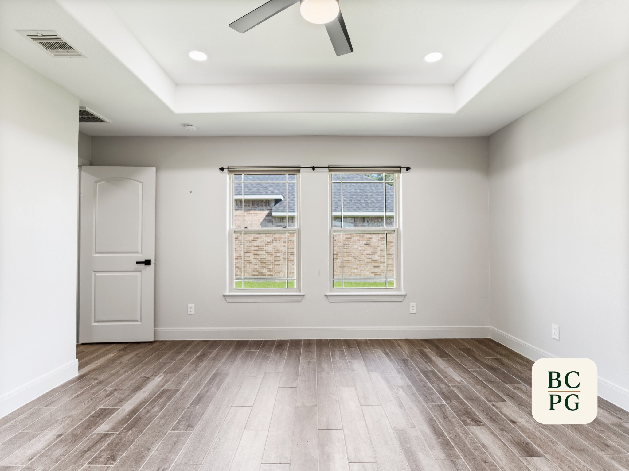 Empty room with white walls, brown wood flooring, two large windows, partially covered by white blinds, white door, ceiling fan, and ceiling lights.