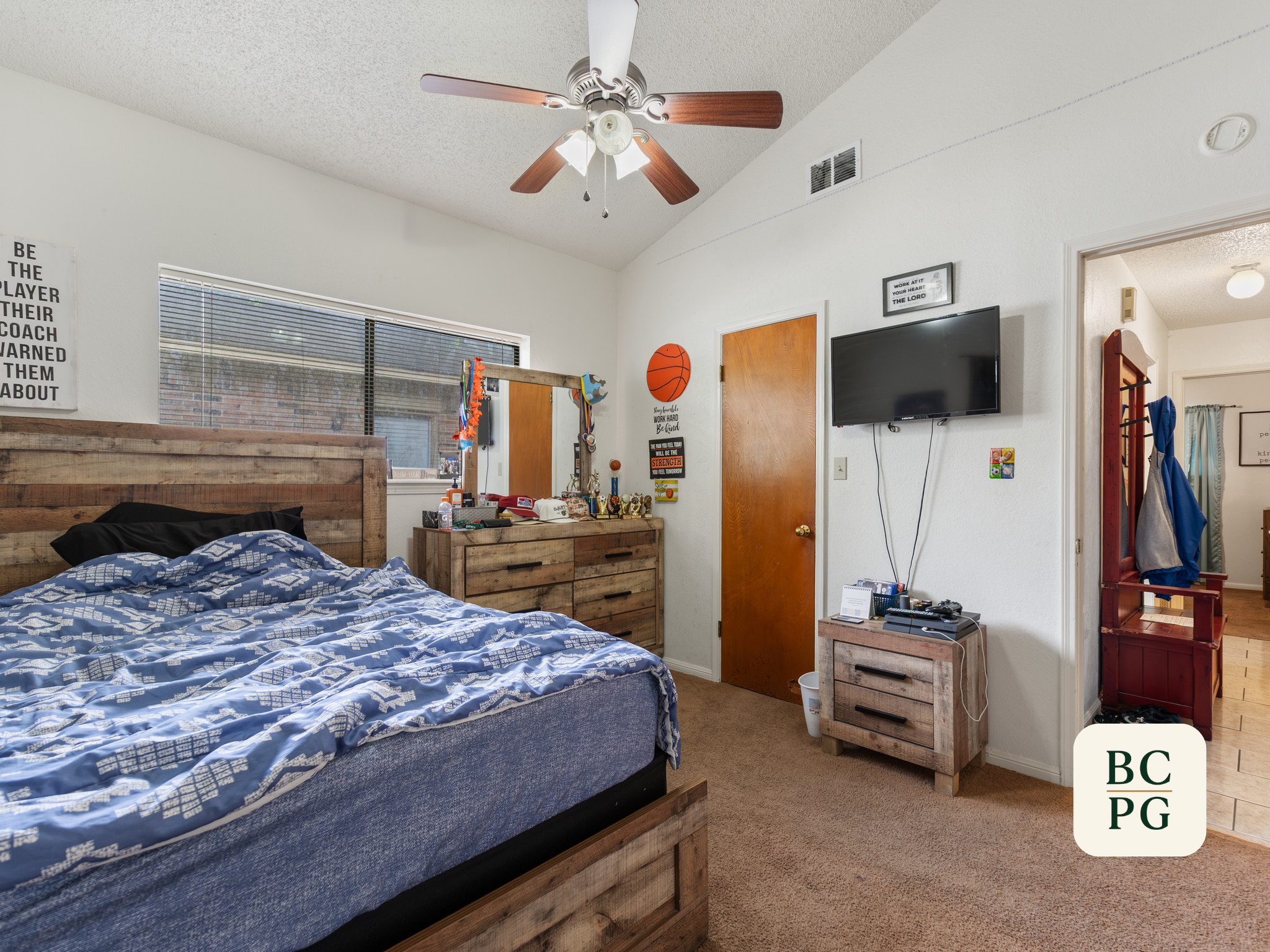 Interior of a bedroom with a wooden bed frame, blue patterned bedding, wooden dresser, wall decorations, a mounted flat-screen TV, ceiling fan, and a view into a bathroom