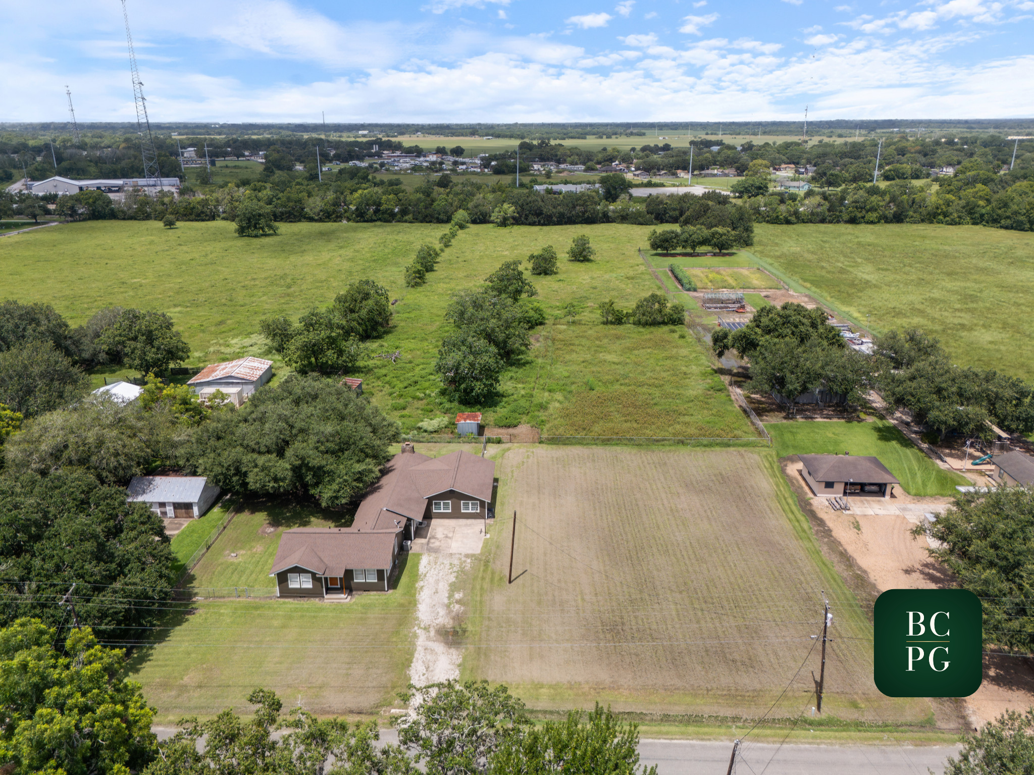 Aerial view of a rural property with a house, garden, and farmland, surrounded by trees and open fields, with a distant town and power lines in the background.