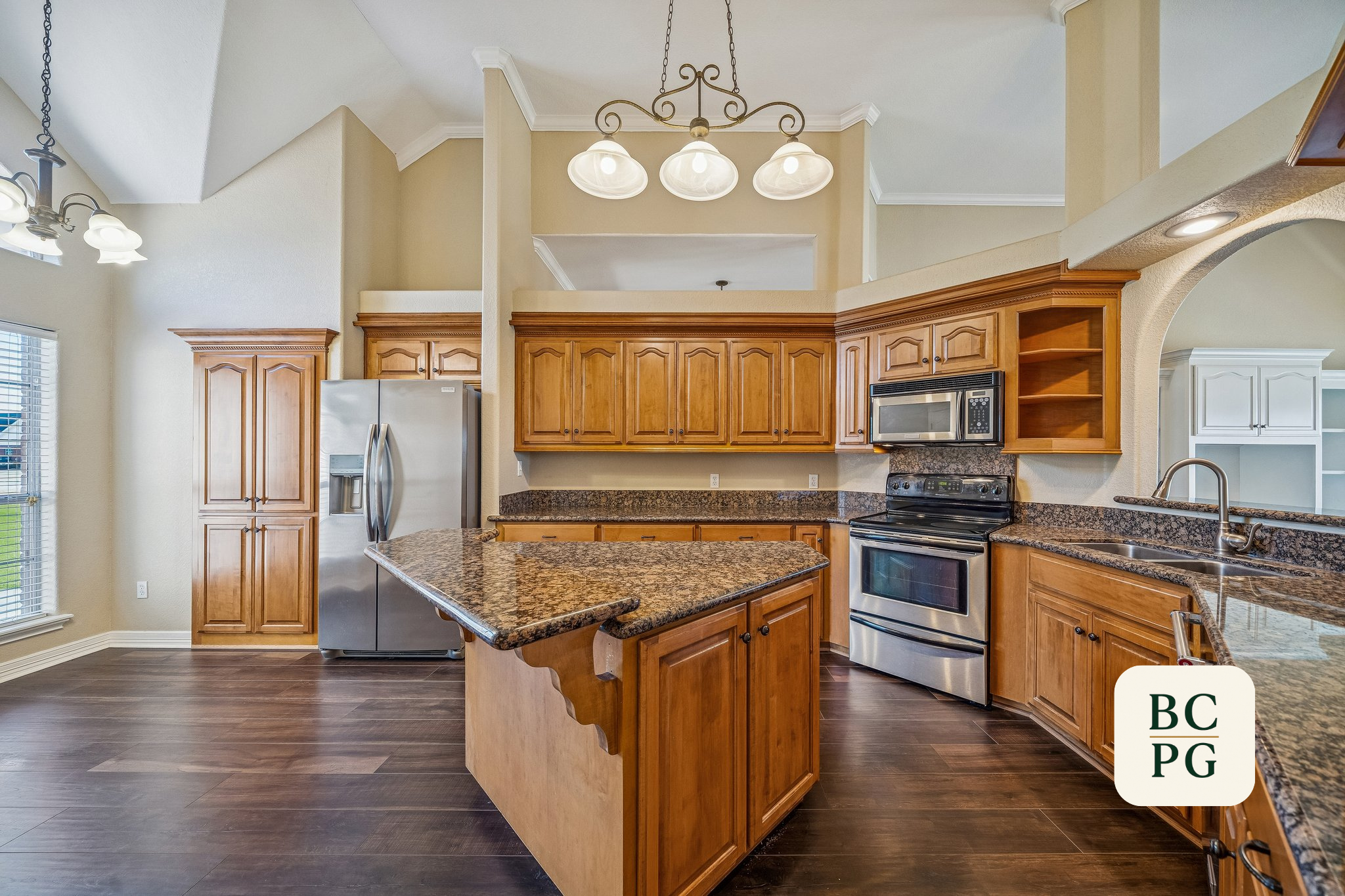 Kitchen with wooden cabinets, granite countertops, stainless steel appliances, a kitchen island, and an archway. Chandeliers hang from the ceiling, and there are windows with blinds on the left wall.