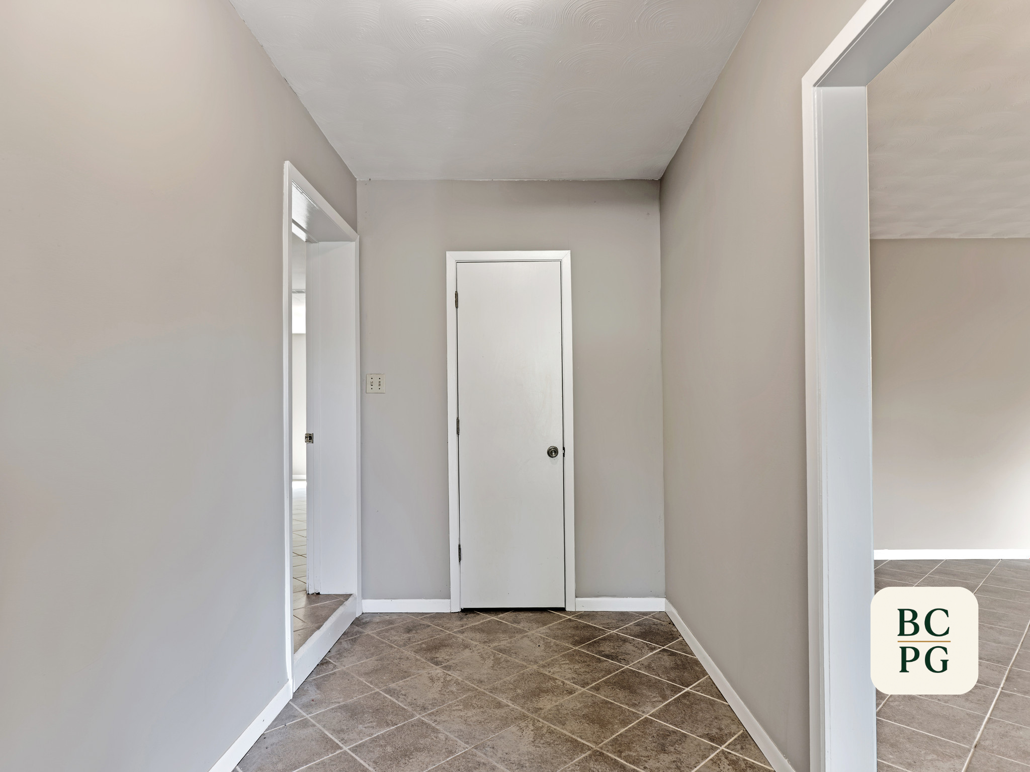 Interior view of a hallway with white walls, a white door at the end, and tiled dark floor. There are wall openings leading to other rooms.