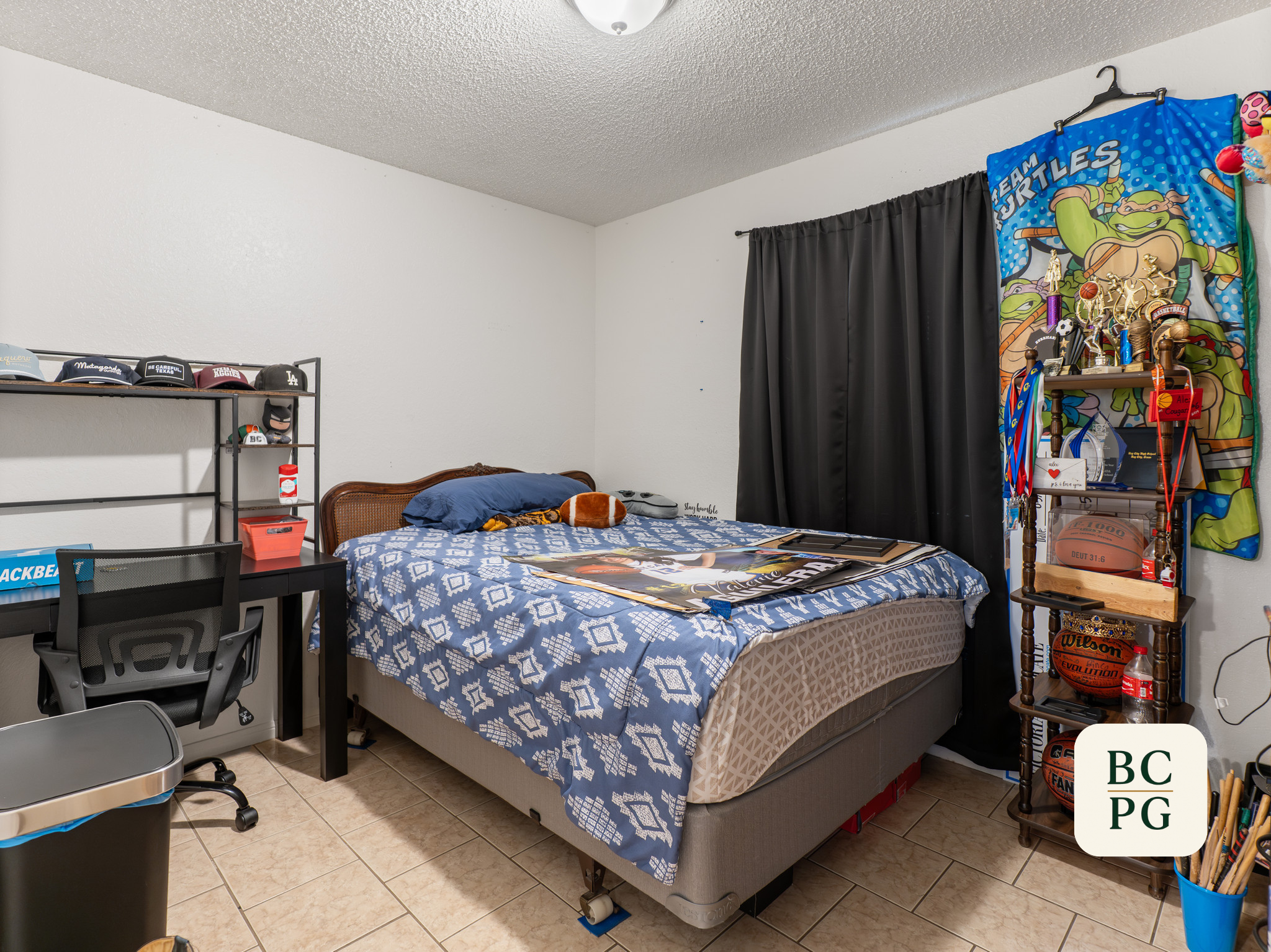 A bedroom with a bed covered in a blue and white patterned bedspread, with pillows and plush toys on it. To the right, there's a wooden shelf with trophies, medals, basketballs, soccer balls, and various items. A black curtain covers a window. On the