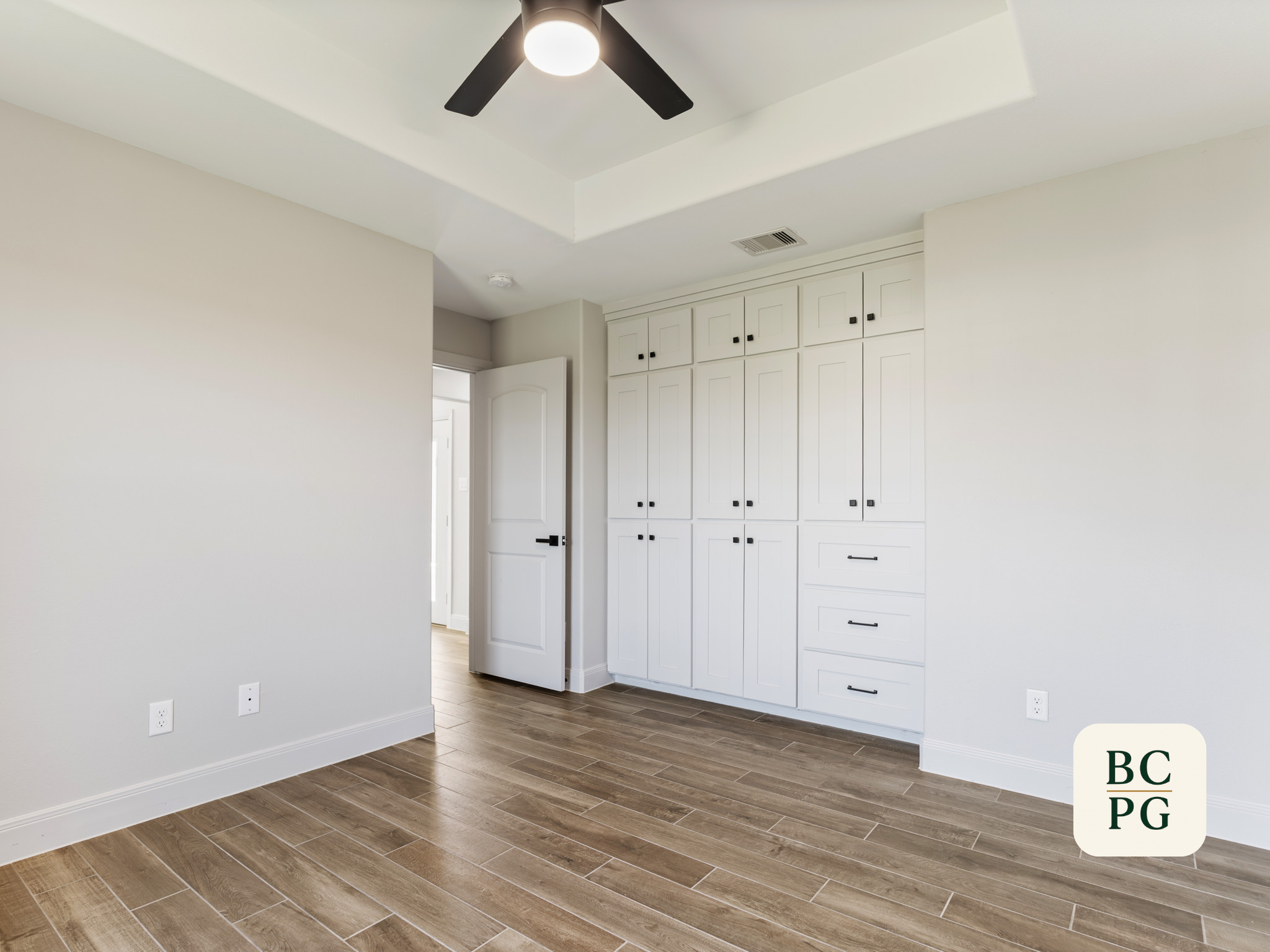 Empty room with white walls, dark wood flooring, a ceiling fan with a light, and built-in white cabinets with black handles.