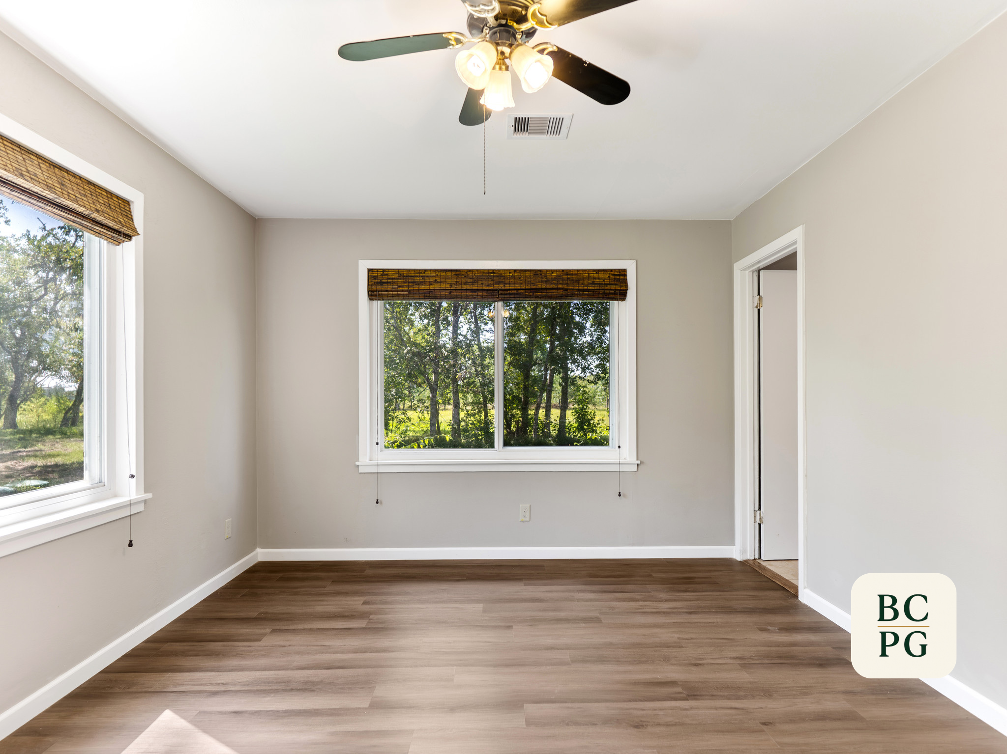 Empty room with two windows, light-colored walls, wooden flooring, ceiling fan with lights, and a door.