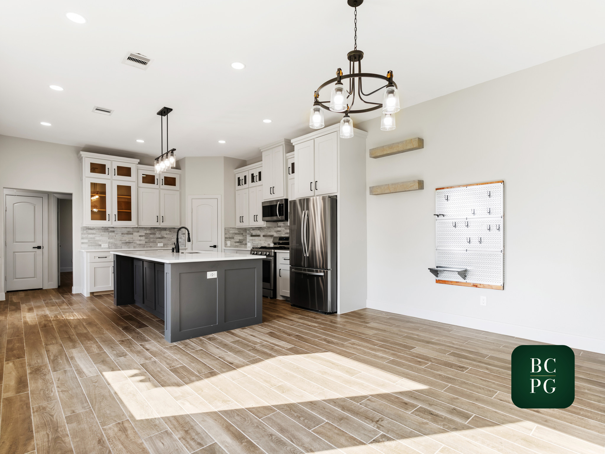 A modern kitchen with white cabinets, stainless steel appliances, a black kitchen island, a tiled backsplash, wood flooring, and two black hanging light fixtures. There are floating shelves on the wall and a pegboard for organizing utensils.