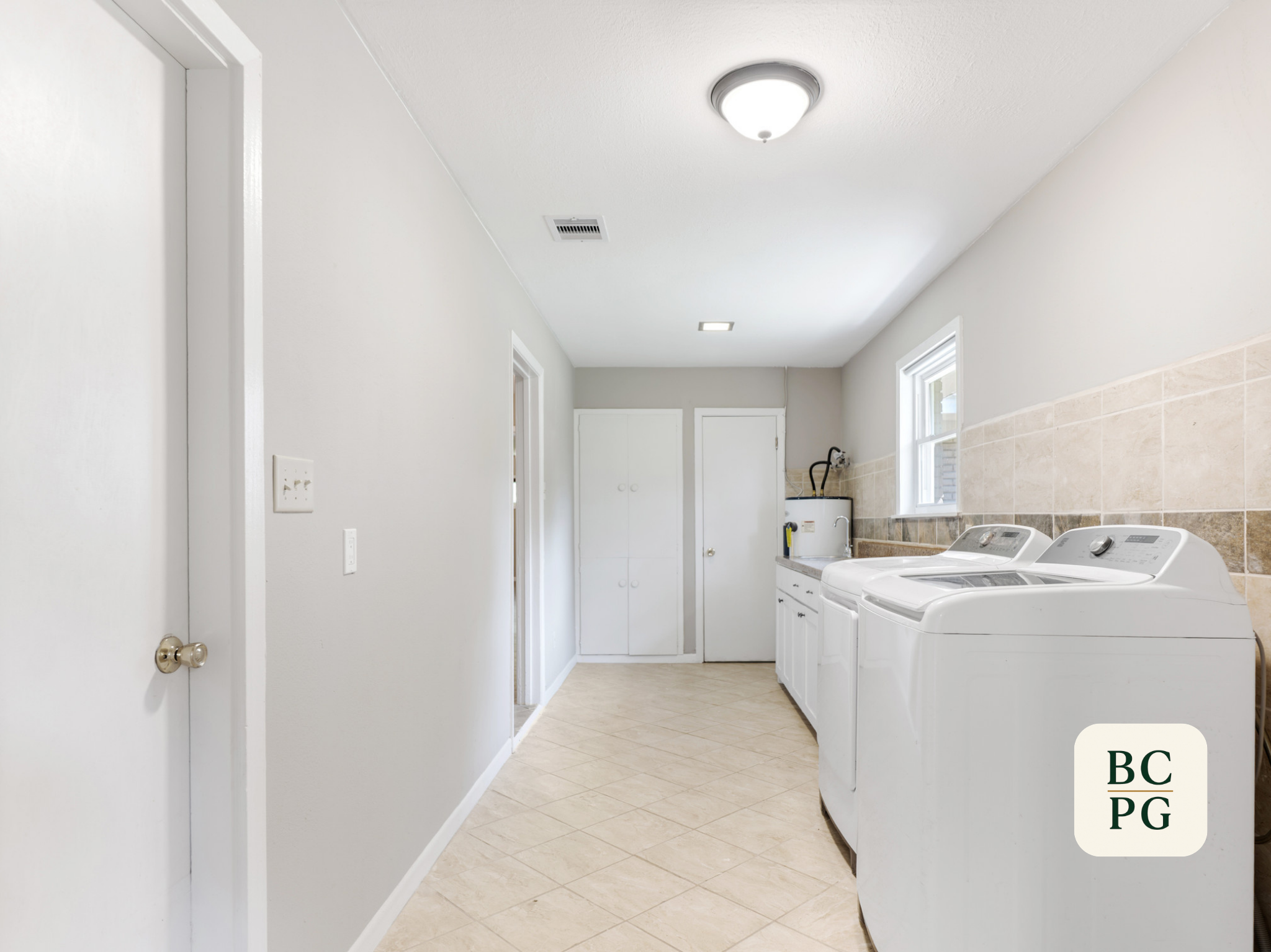 A laundry room with a white washing machine and dryer, beige tiled floor, white walls, a small window, and a ceiling light fixture.