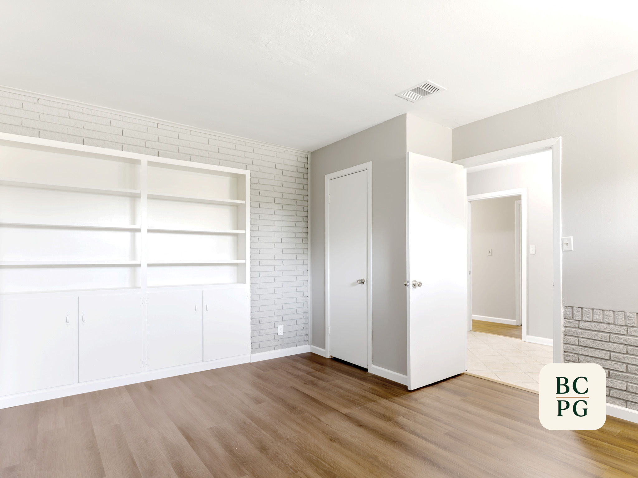 Interior view of a living room with white built-in shelves, white brick wall, and open doorway leading to a kitchen with tiled floor and wooden flooring in the living room.