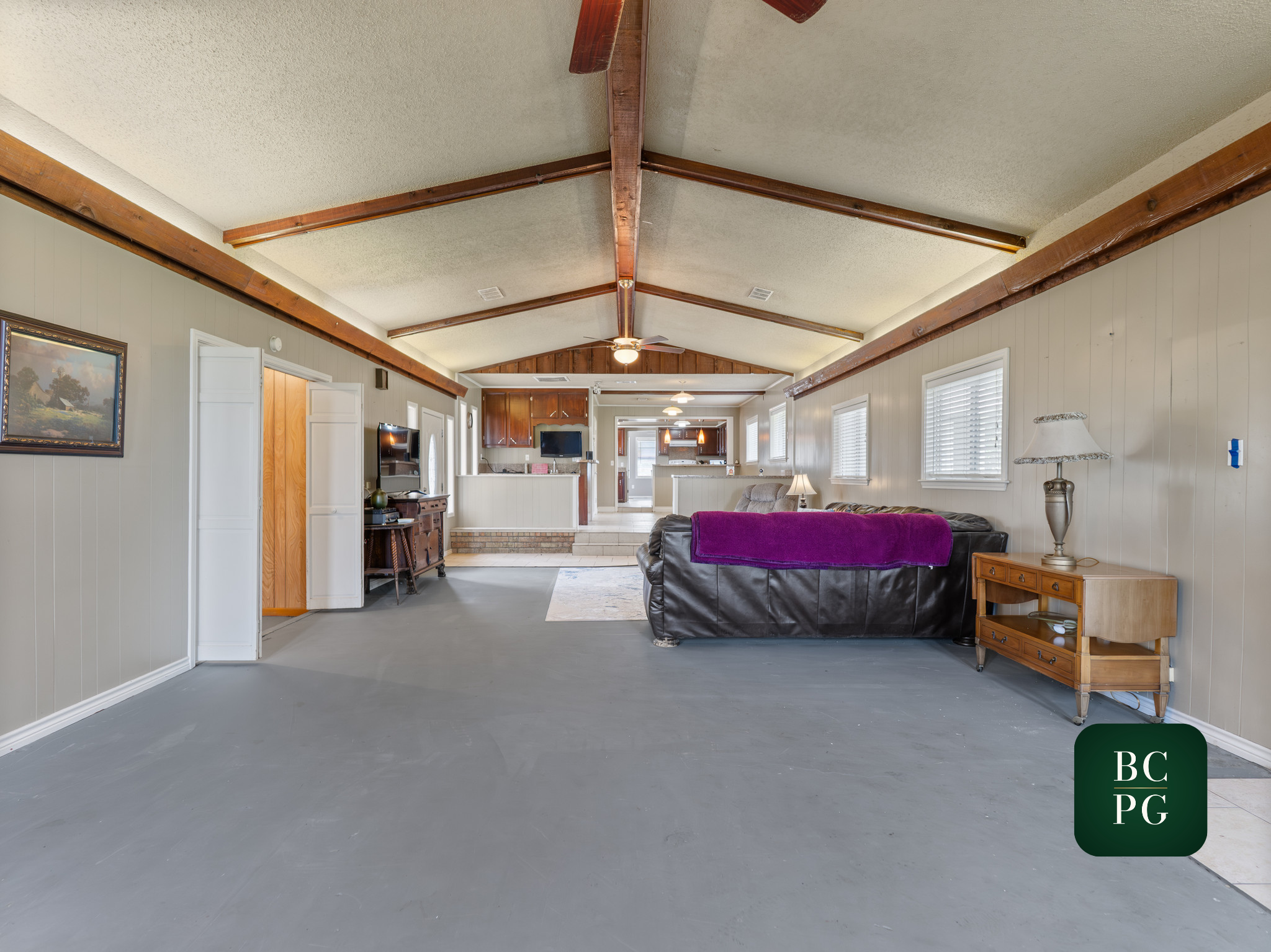 Open-concept living room with vaulted ceiling featuring wooden beams, built-in shelves, and large windows, with furniture including a TV, armchair, and sofa.