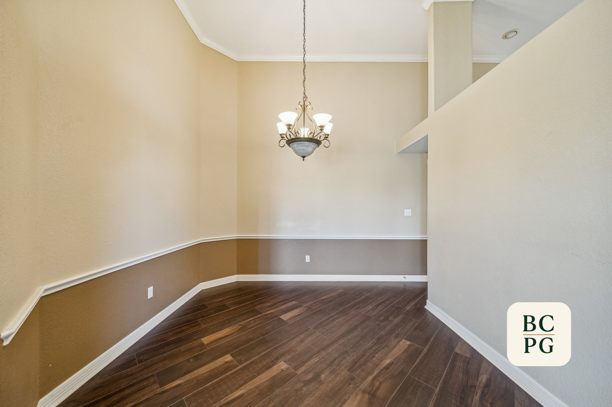 Empty interior space with beige and brown walls, dark hardwood floor, chandelier hanging from ceiling, and corner with an electrical outlet.