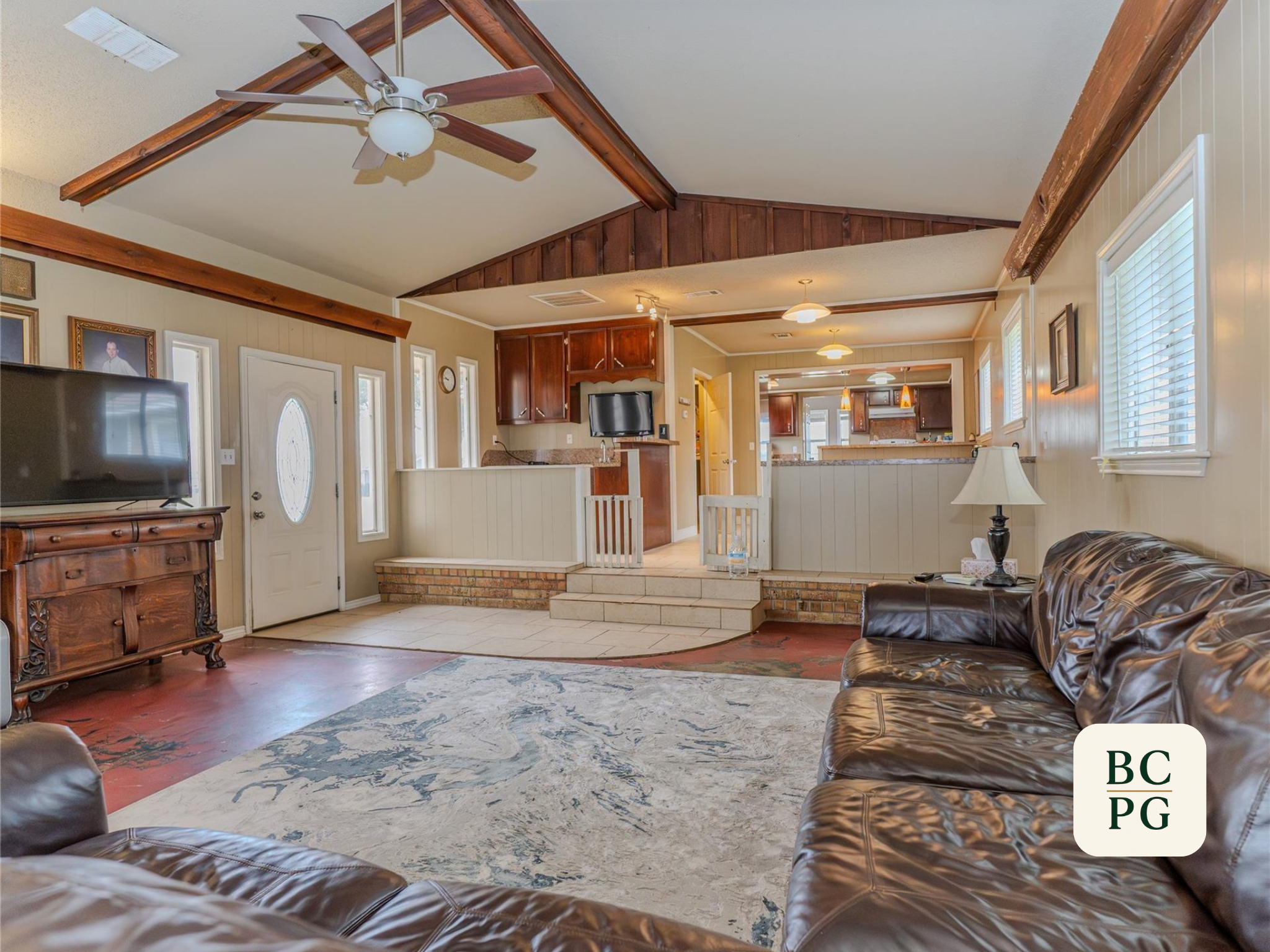 Interior of a living room with ceilings featuring exposed wooden beams, a ceiling fan, and multiple windows allowing natural light. There is a dark leather sofa, a TV mounted on the wall, and a patterned rug on the floor. The room opens into a kitche