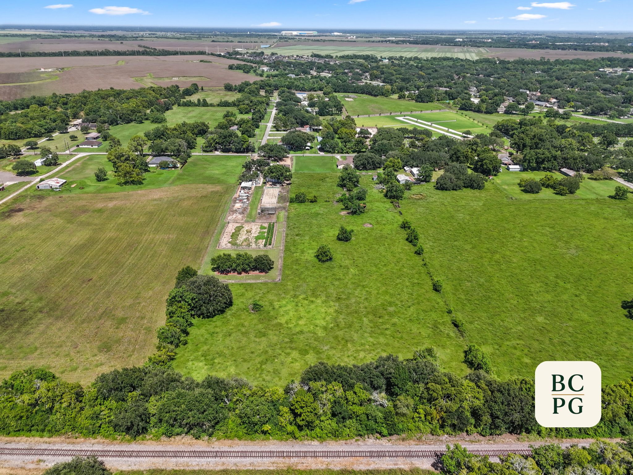 Aerial view of a rural landscape with houses, green lawns, trees, and farmland, with a railroad track in the foreground and a cityscape in the distant background.