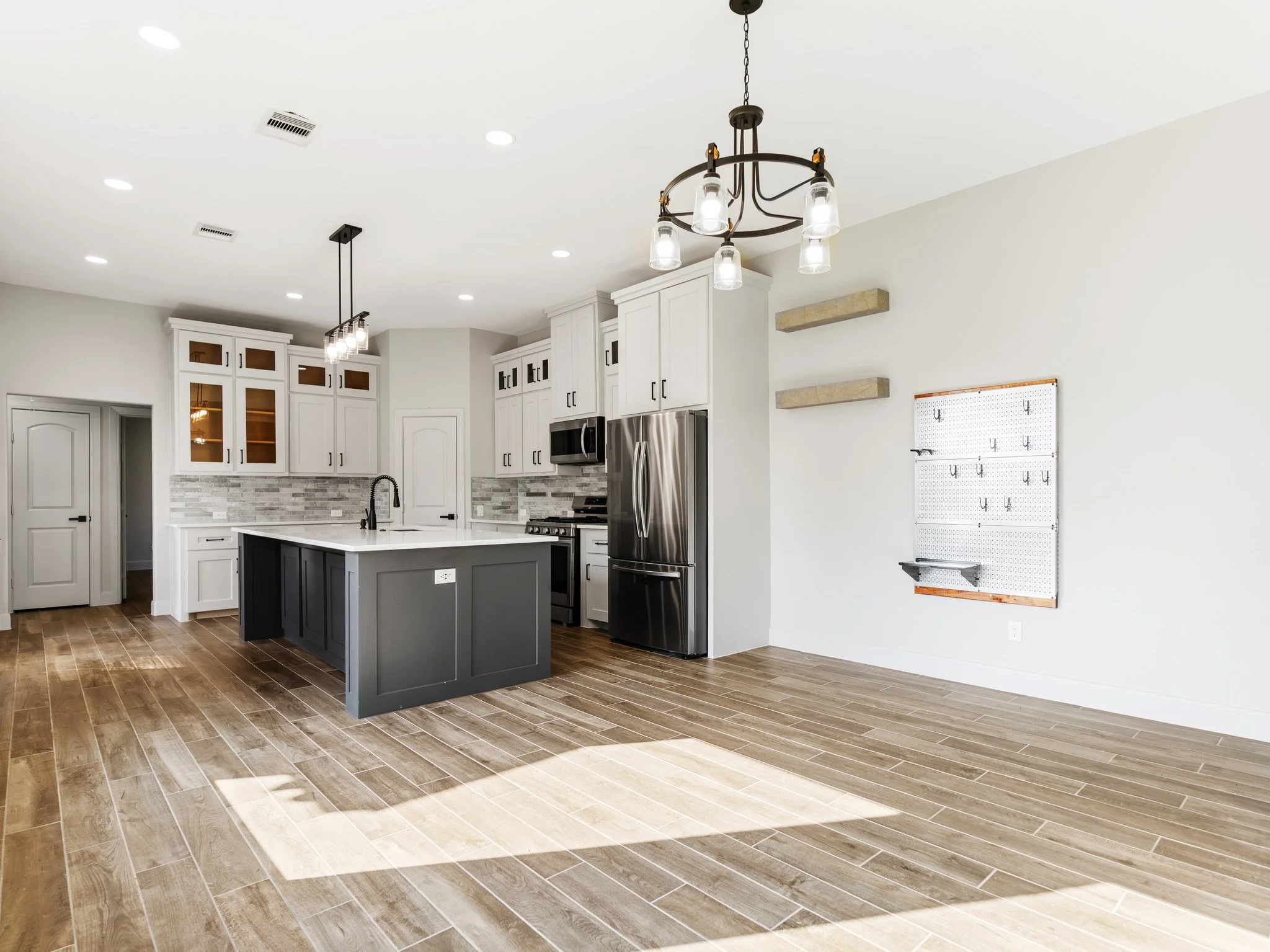 Empty modern kitchen with white cabinets, gray island, stainless steel refrigerator, and wood flooring, featuring two hanging light fixtures and a pegboard on the wall.