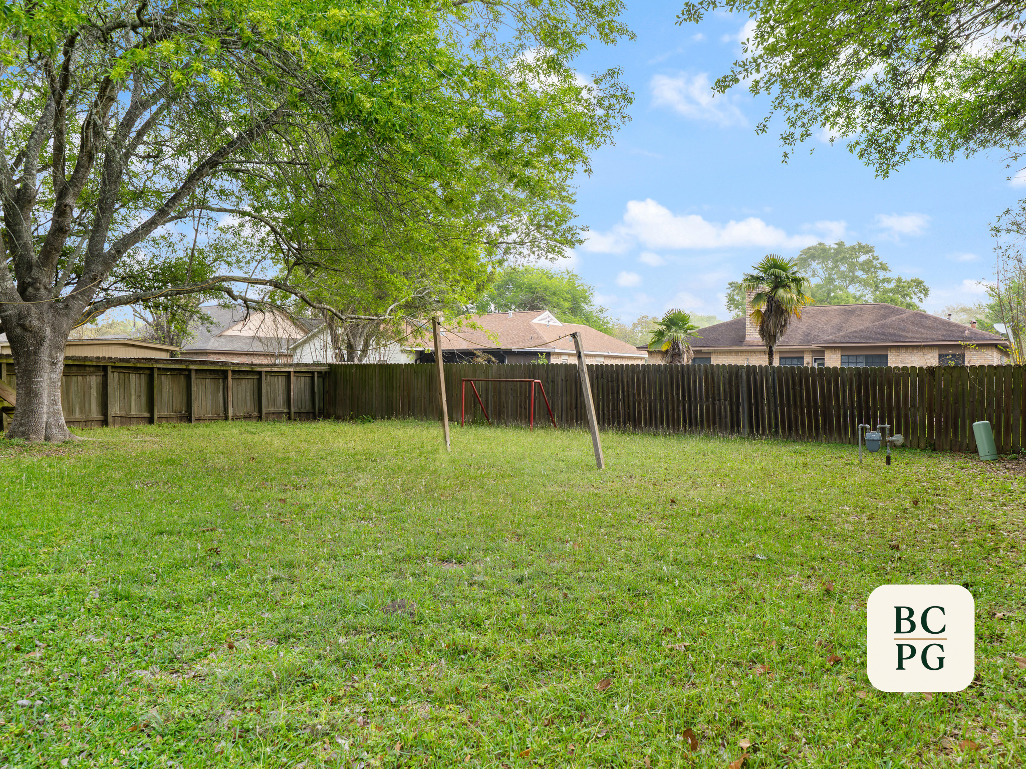 Backyard with large leafy tree, wooden fence, and houses in the background under a partly cloudy sky.