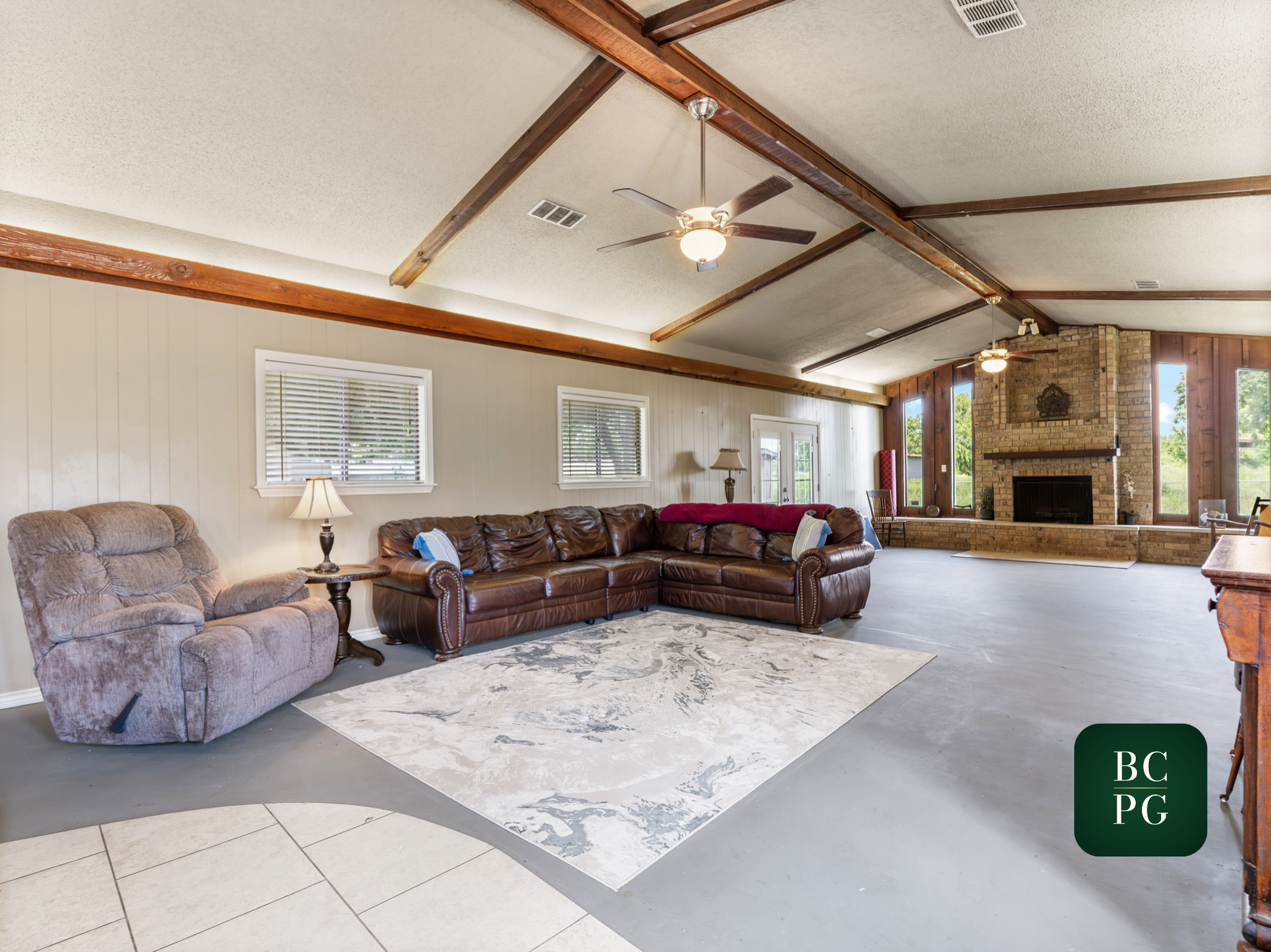 Living room with a vaulted ceiling having wooden beams, ceiling fans, a brick fireplace, large windows, and various furniture including a brown leather sofa and an armchair.