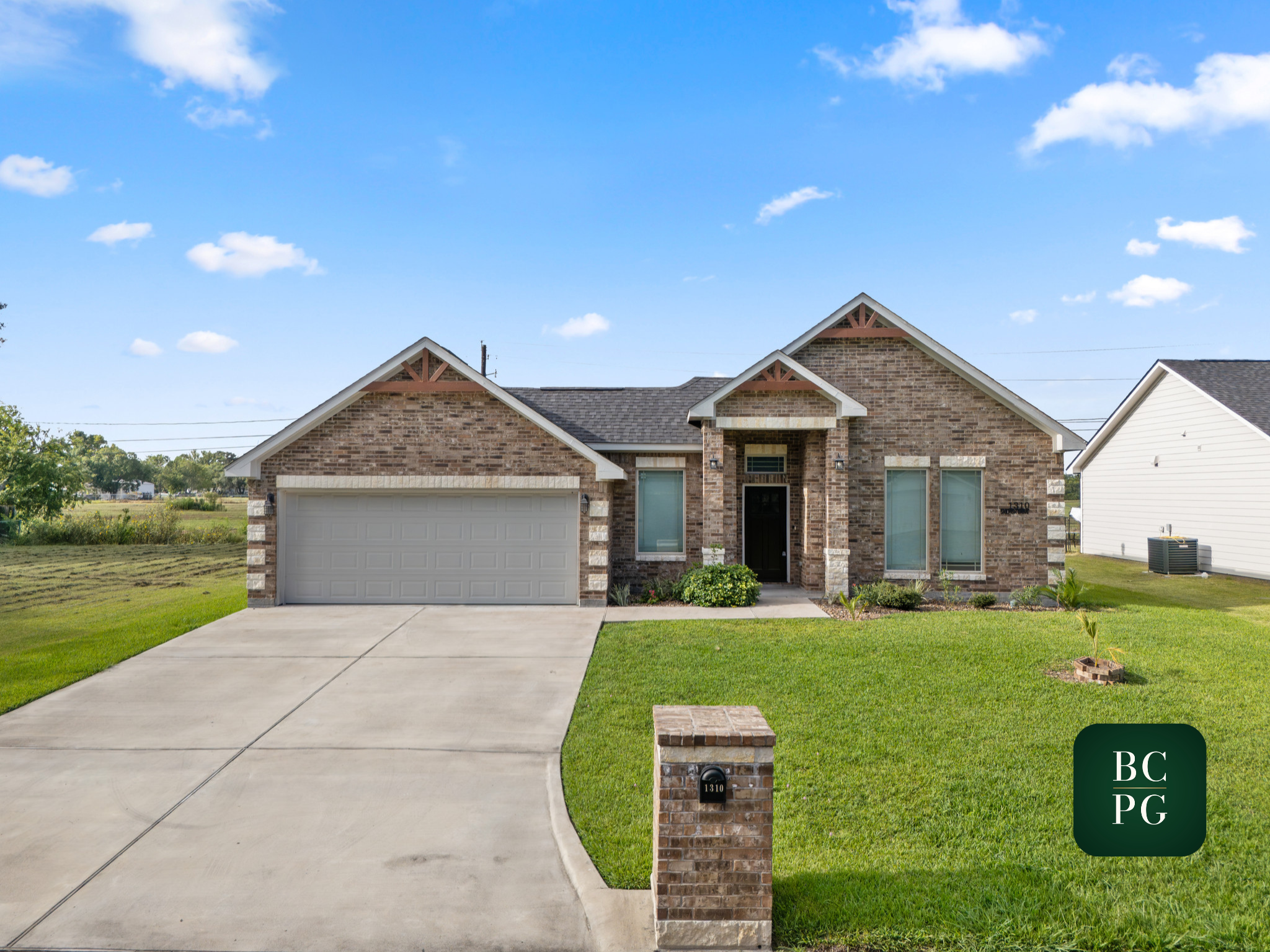 Front view of a brick house with a two-car garage, a front porch, and a green lawn, under a blue sky with clouds.