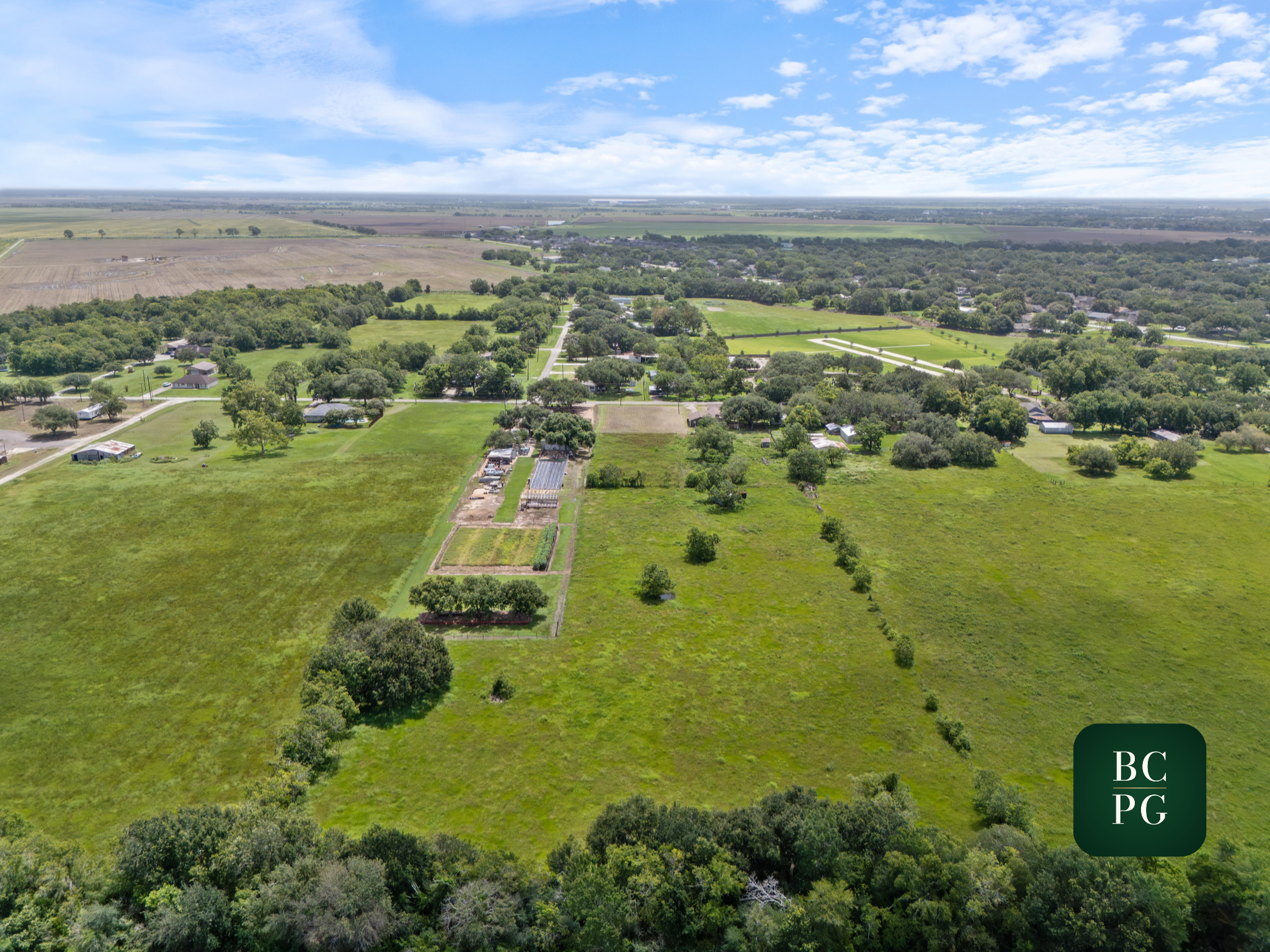 Aerial view of a rural landscape with green fields, trees, and scattered houses under a partly cloudy sky.