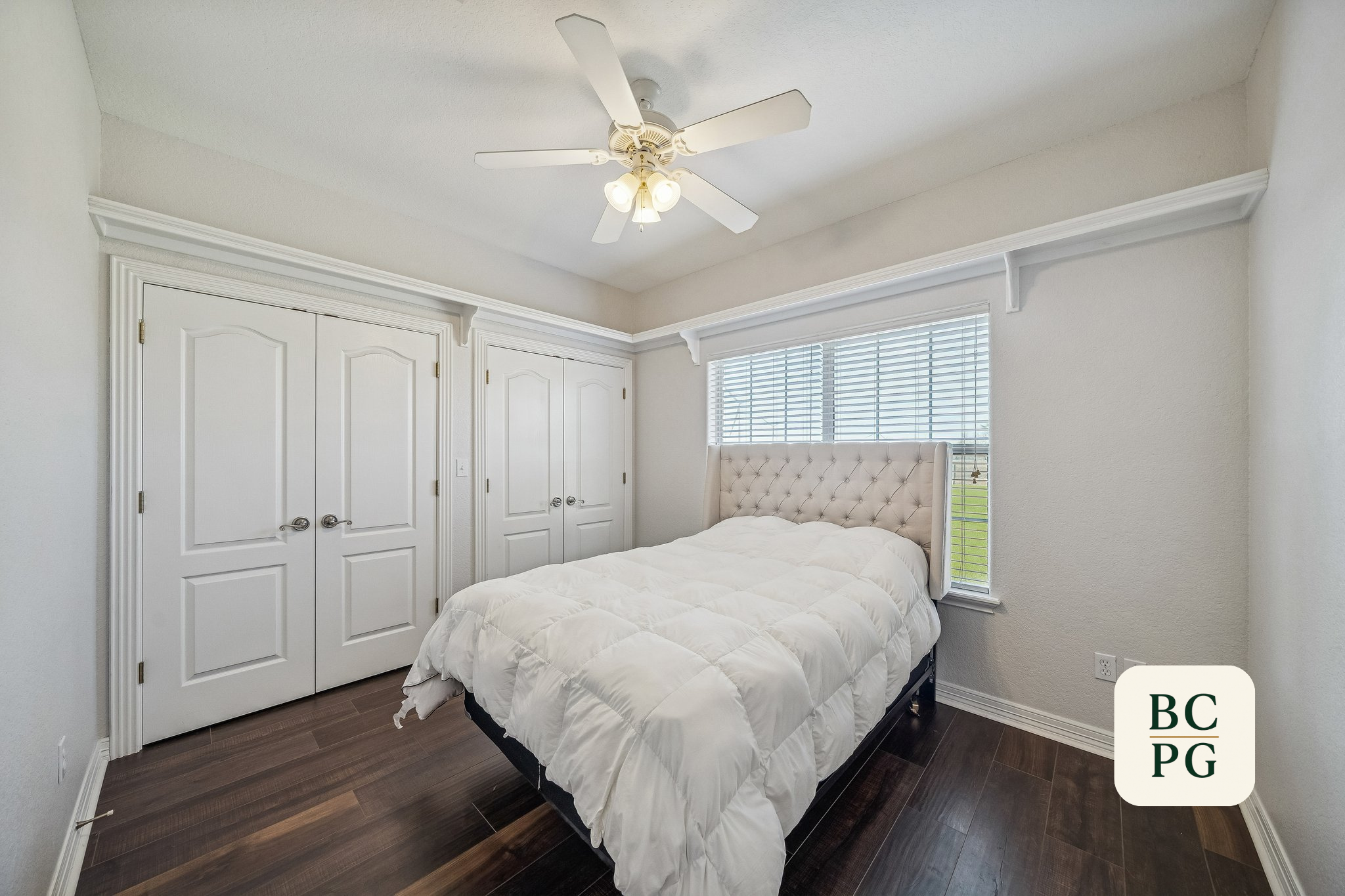 A bedroom with a white ceiling fan, white walls, closed white closet doors, a window with blinds, a bed with a white comforter and tufted headboard on dark wood flooring.