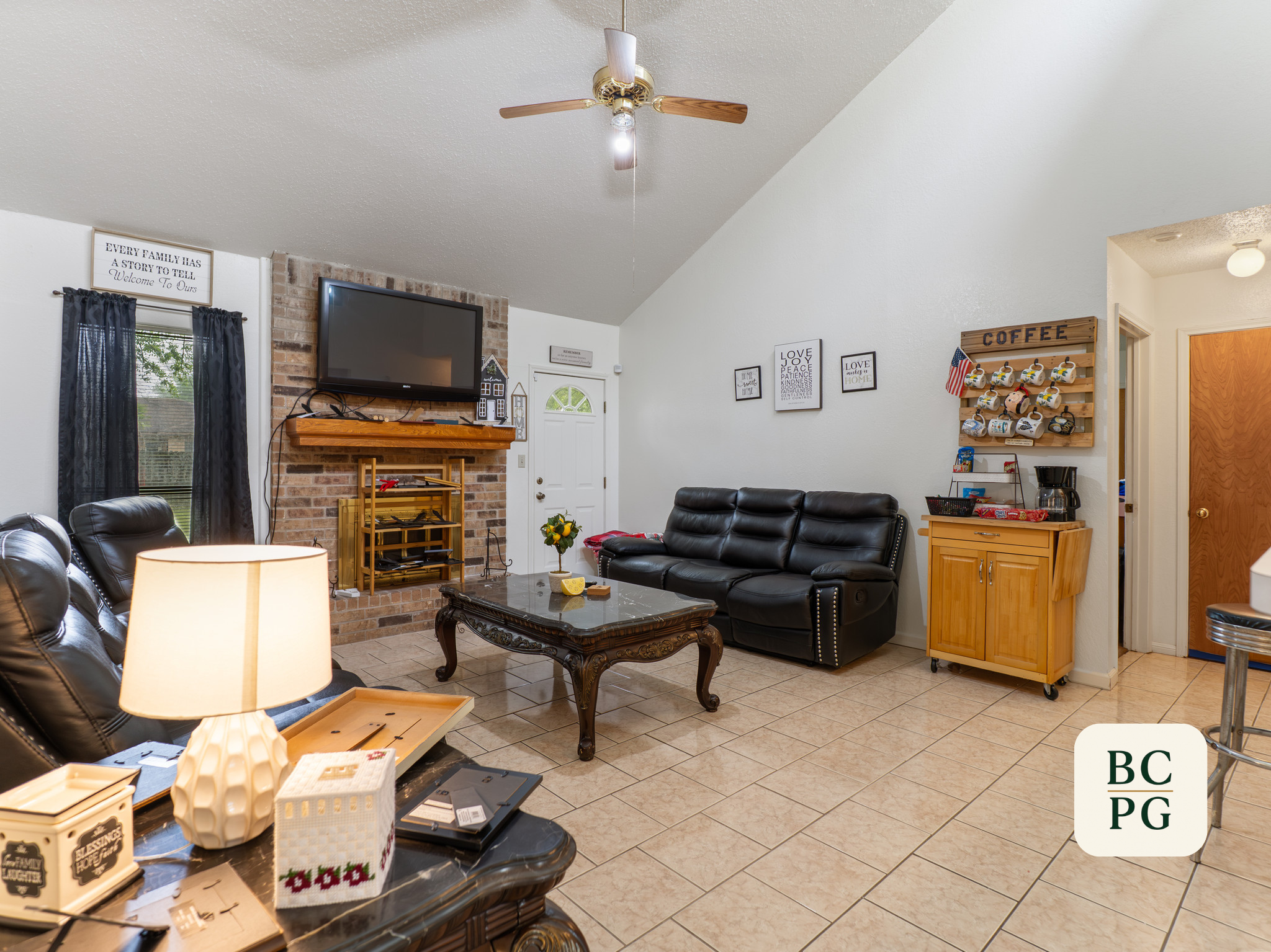 Living room with a black leather sofa, a coffee table, and various decorations including framed quotes and a wall-mounted flat-screen TV. There is a ceiling fan with wooden blades, a brick fireplace, and a window with dark curtains.