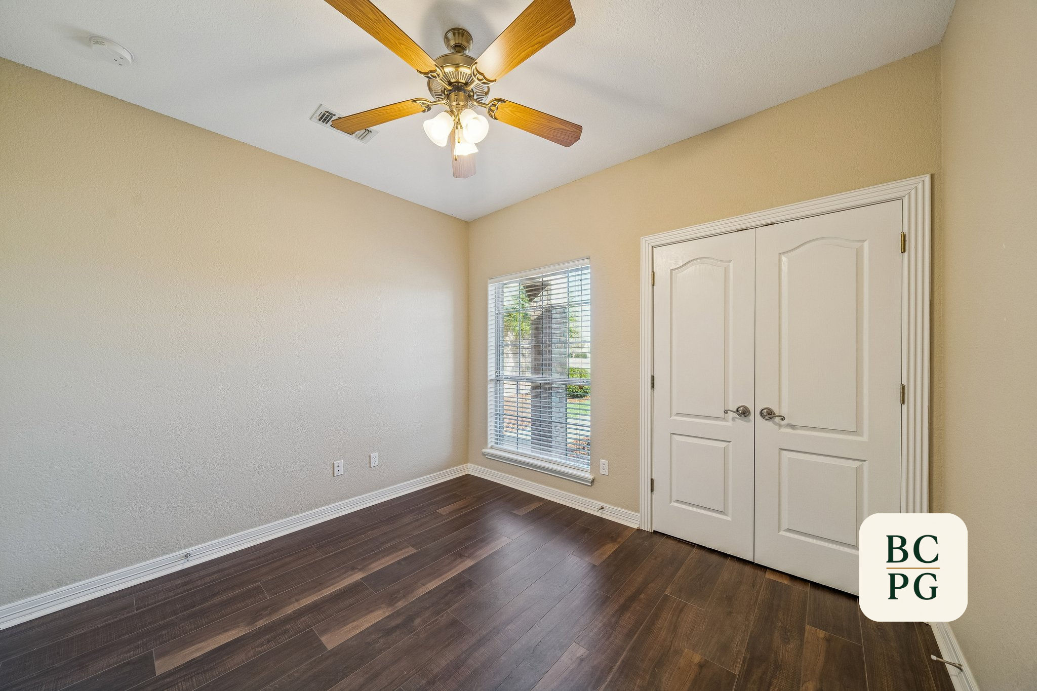 Empty room with beige walls, hardwood flooring, a white double door closet, a window with blinds showing an outside view, and a ceiling fan with wooden blades and a light fixture.