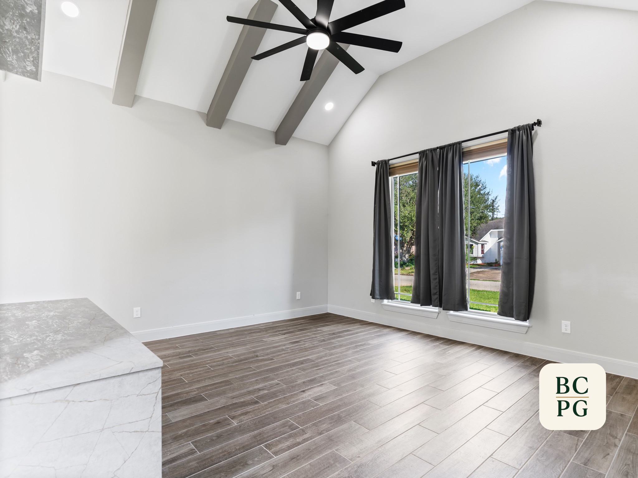 Interior of a modern house with a high ceiling, black ceiling fan, large window with gray curtains, white walls, and wood-look tile flooring