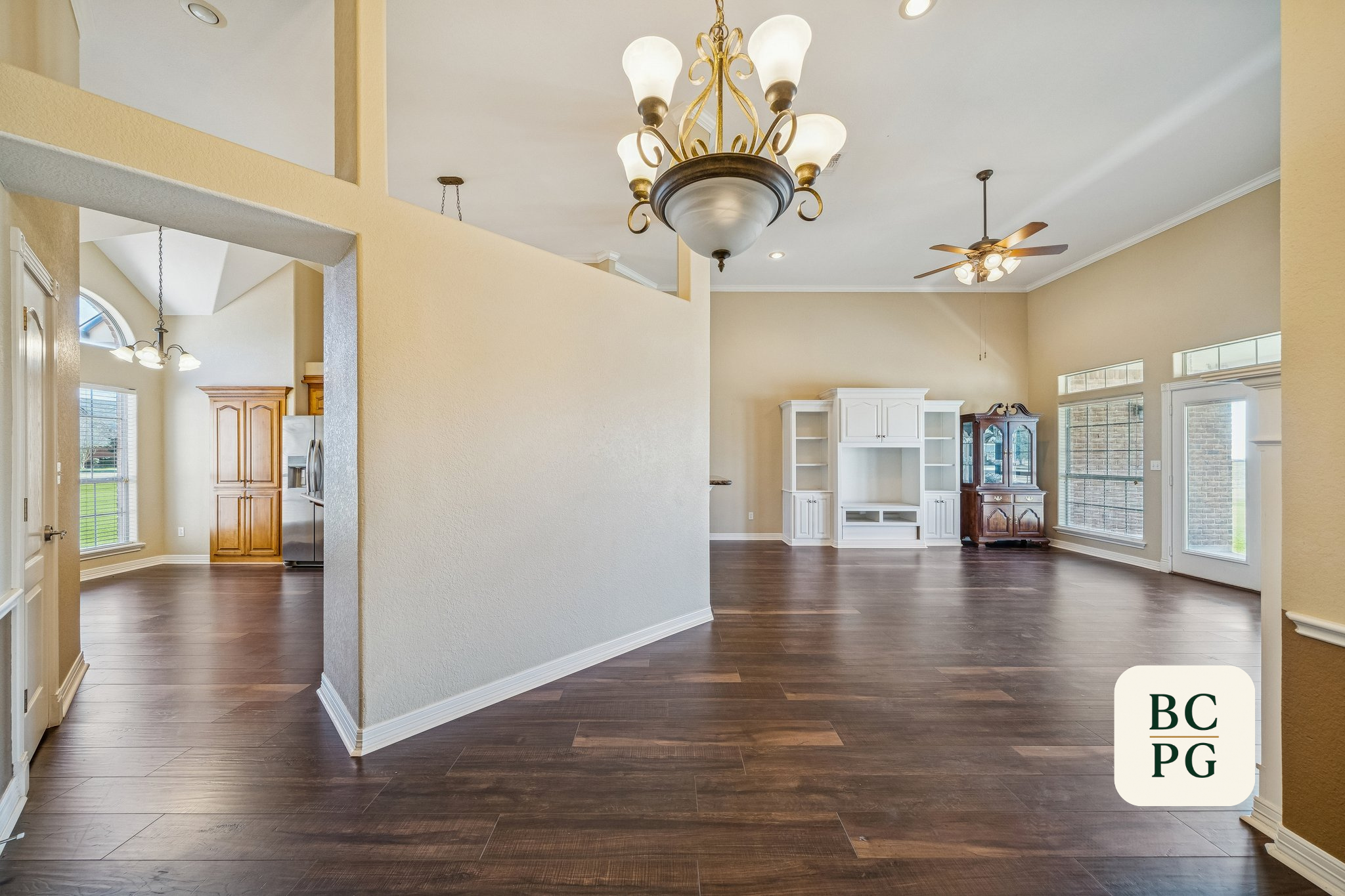 Living room with dark wood flooring, beige walls, large windows, white built-in shelf, and various light fixtures including a chandelier and ceiling fan.