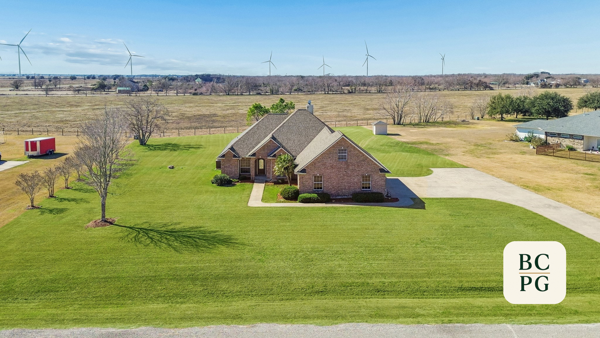 A house with a brick exterior and a chimney, surrounded by a well-maintained green lawn, in a rural area with trees and wind turbines in the background.