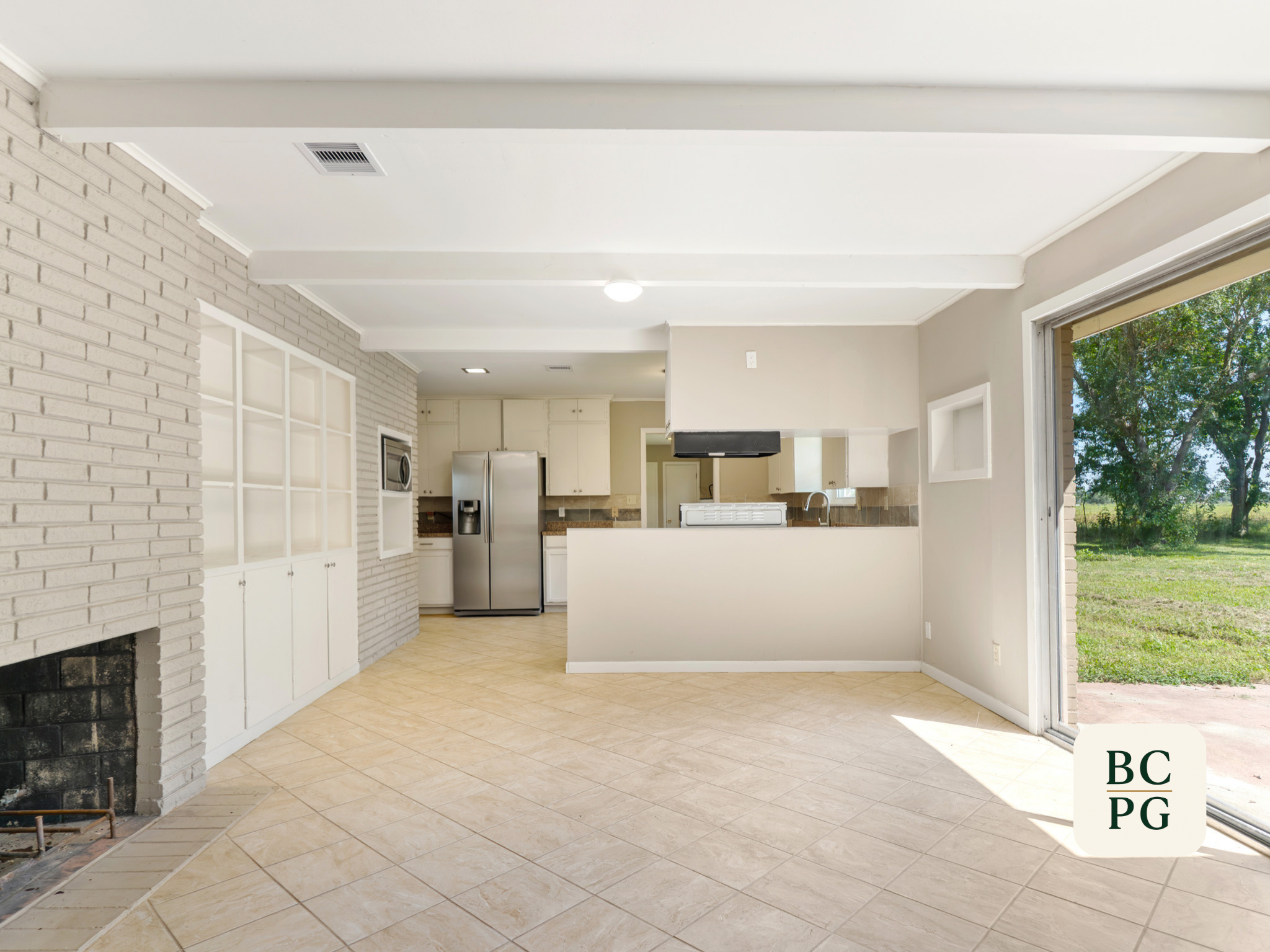 Empty kitchen with white cabinets, stainless steel refrigerator, and large window showing greenery outside.