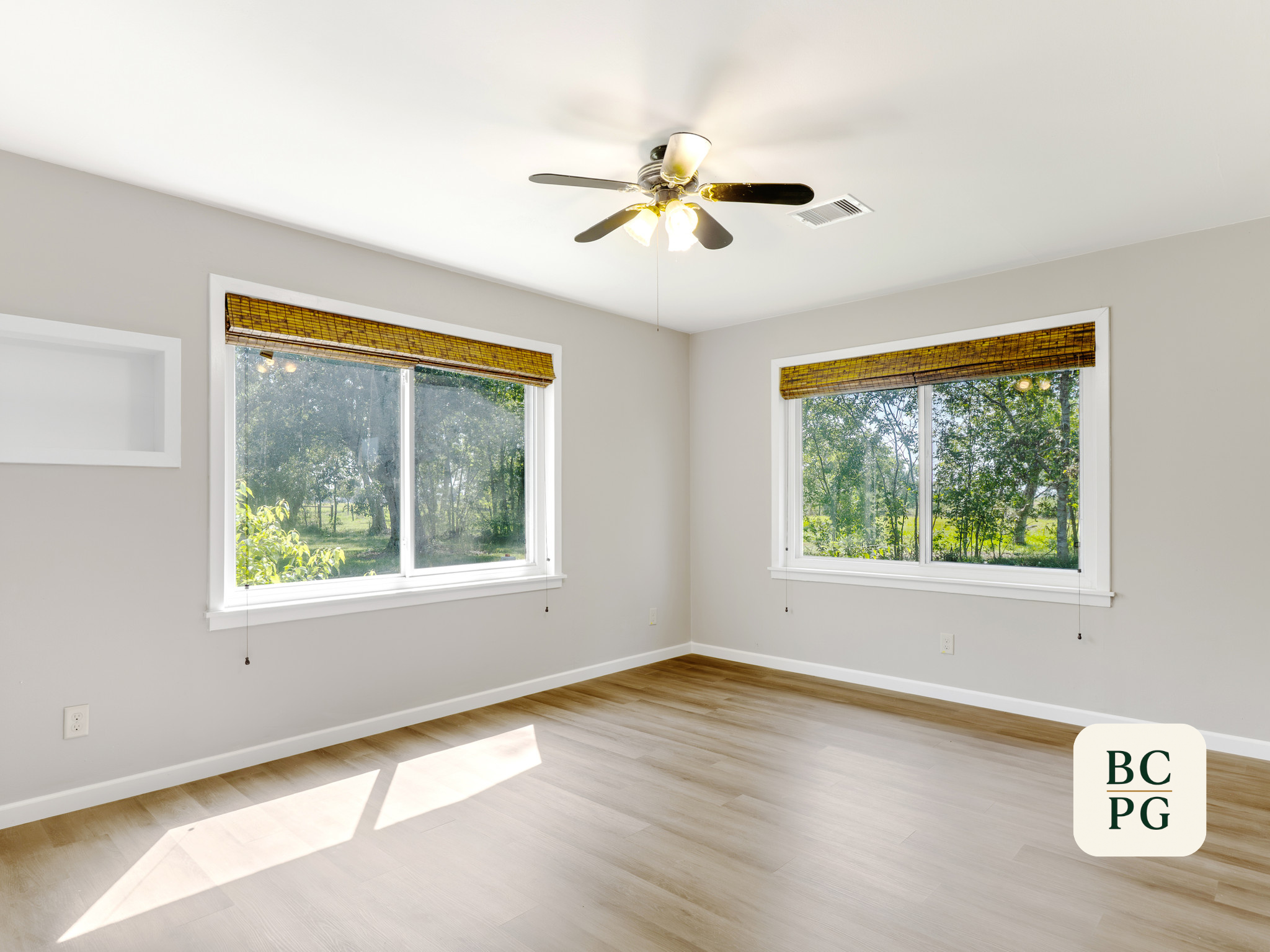 Empty room with large windows, light-colored wooden floor, white walls, and a ceiling fan with attached light fixtures.