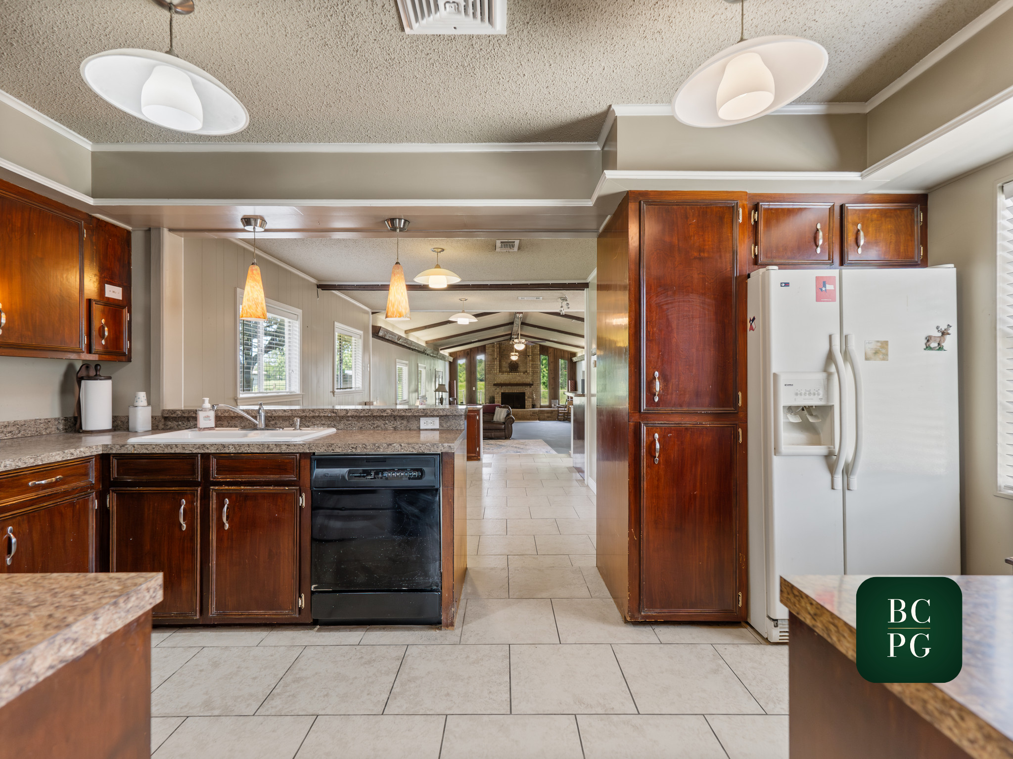 View of a kitchen with wooden cabinets, beige tiled floor, and a glimpse into a living room with a brick fireplace and ceiling fans.