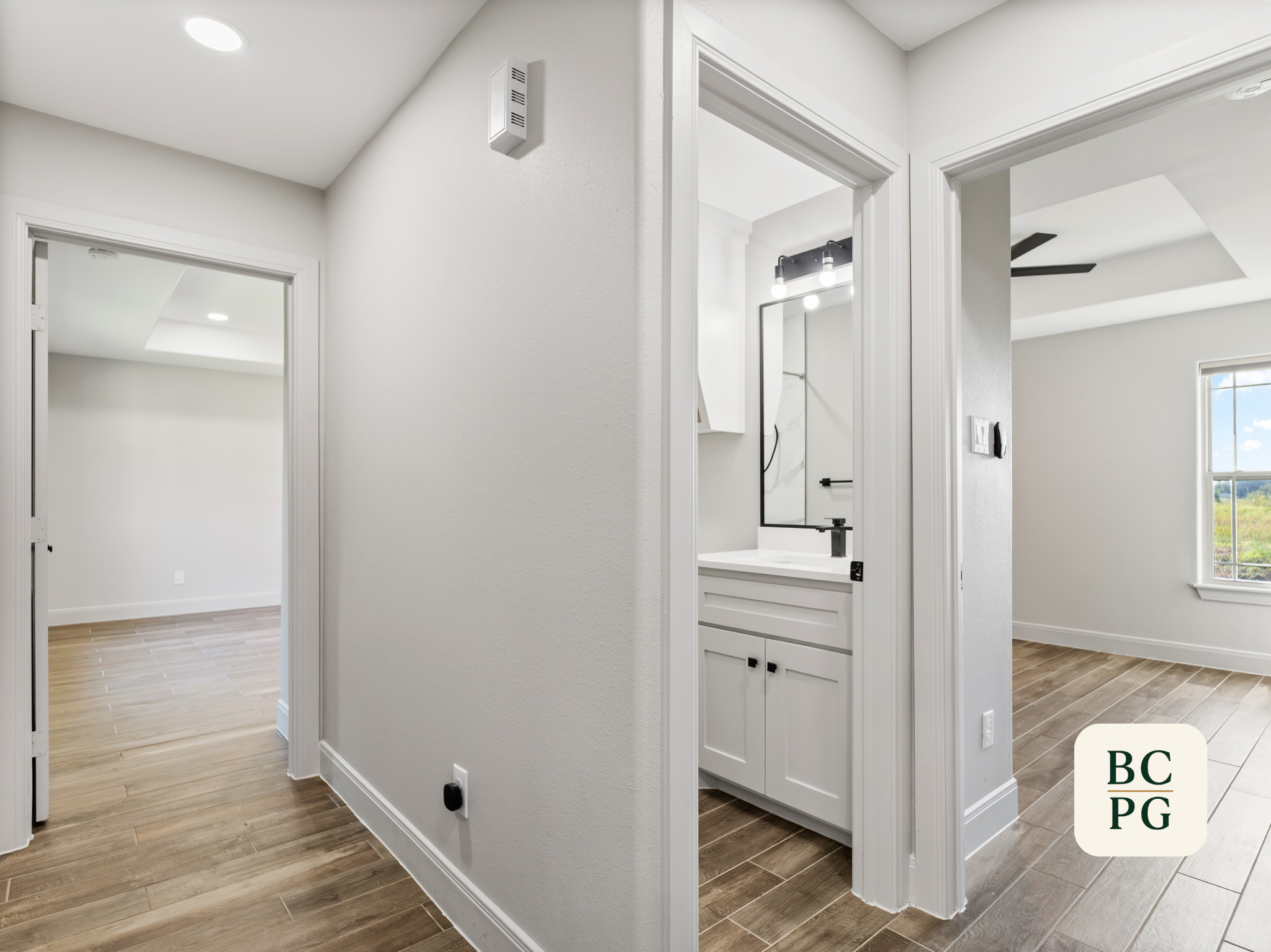 Interior view of a modern home with white walls, wood flooring, and open doorways leading to other rooms. A bathroom with a black light fixture and a mirror is visible.