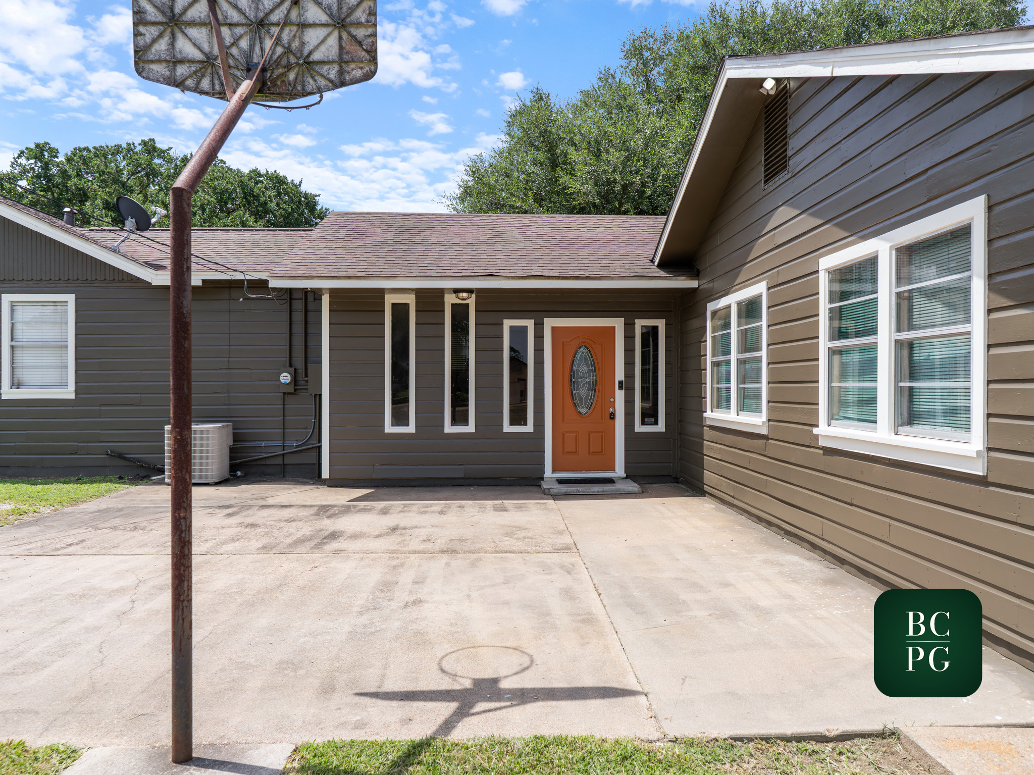 House with a brown shingle roof, gray siding, orange front door, and multiple white-framed windows. There is an old rusted basketball hoop in the foreground, and trees behind the house against a partly cloudy blue sky.