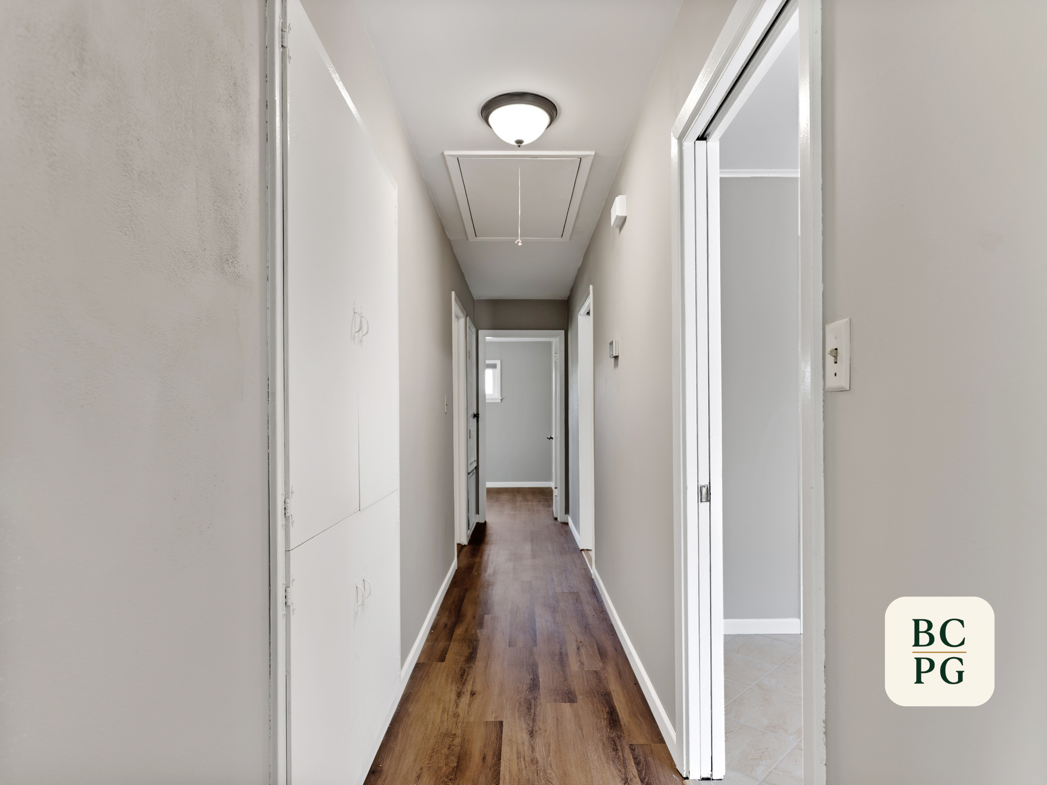 A hallway with white walls, wooden flooring, and multiple doorways leading to different rooms. The ceiling has a flush light fixture and a ceiling hatch with a pull cord.