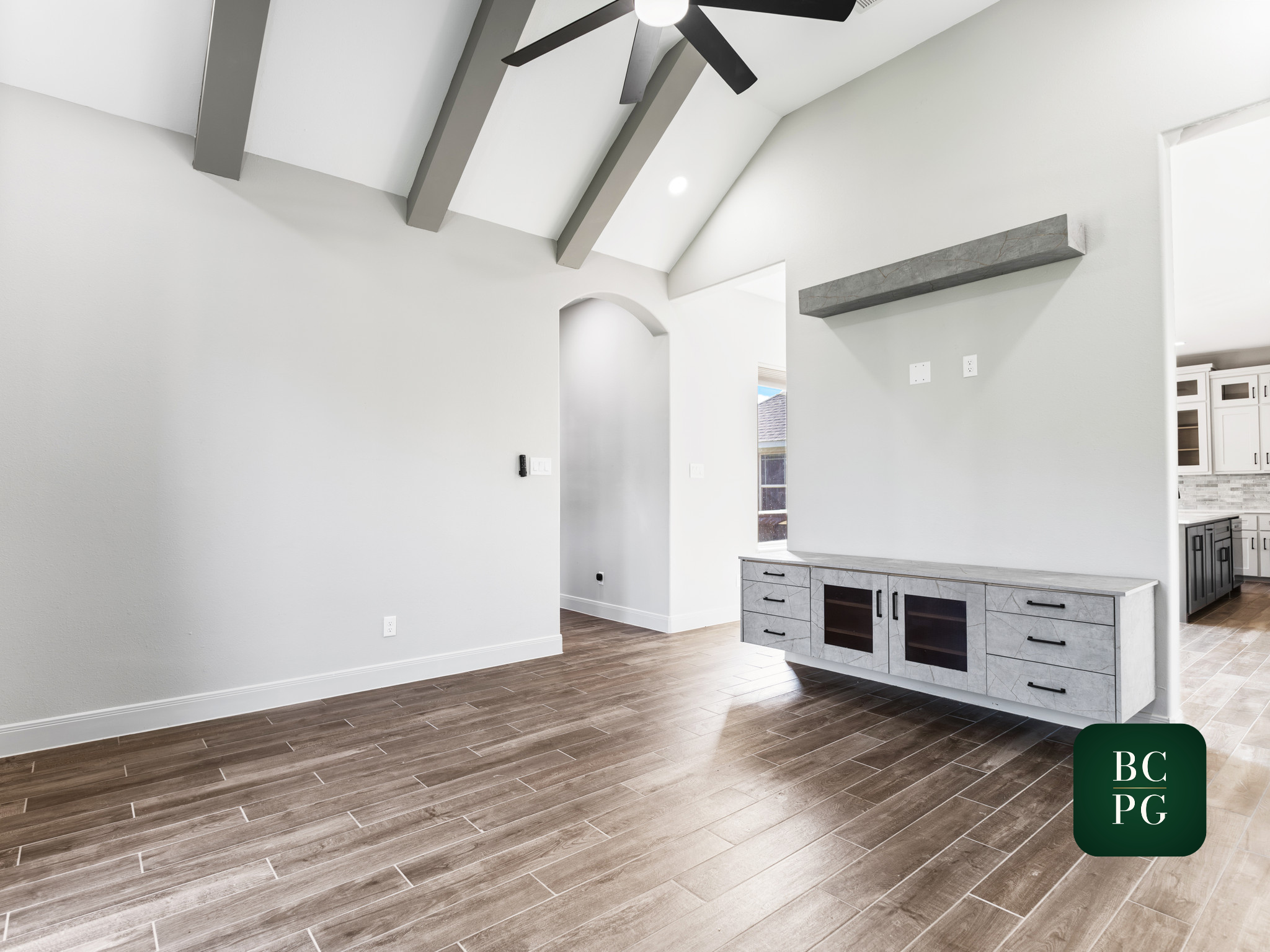 Interior view of a modern, bright living space with white walls, a brown wood floor, a white ceiling with black ceiling fan, and a kitchen area with gray marble cabinets and a window.