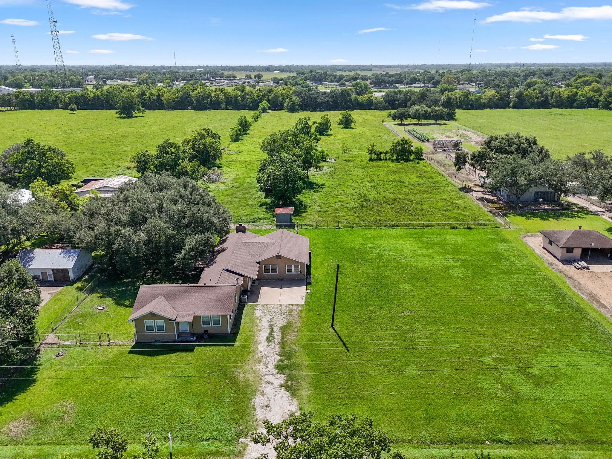 Aerial view of a countryside property with a house, large green fields, trees, and small structures under a clear blue sky.