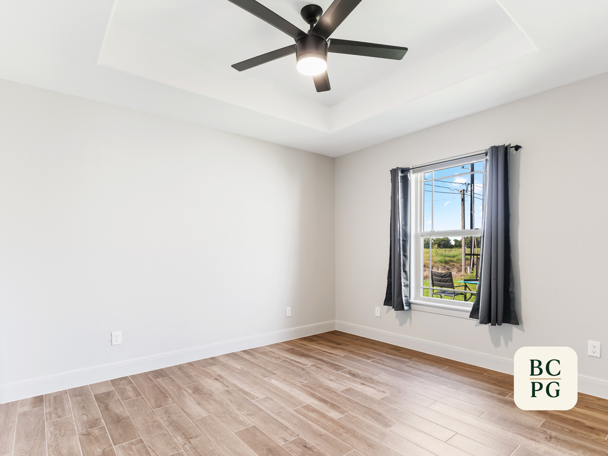 Empty room with white walls, a ceiling fan with a light in the center, a window with dark curtains showing an outdoor view, and wood flooring.