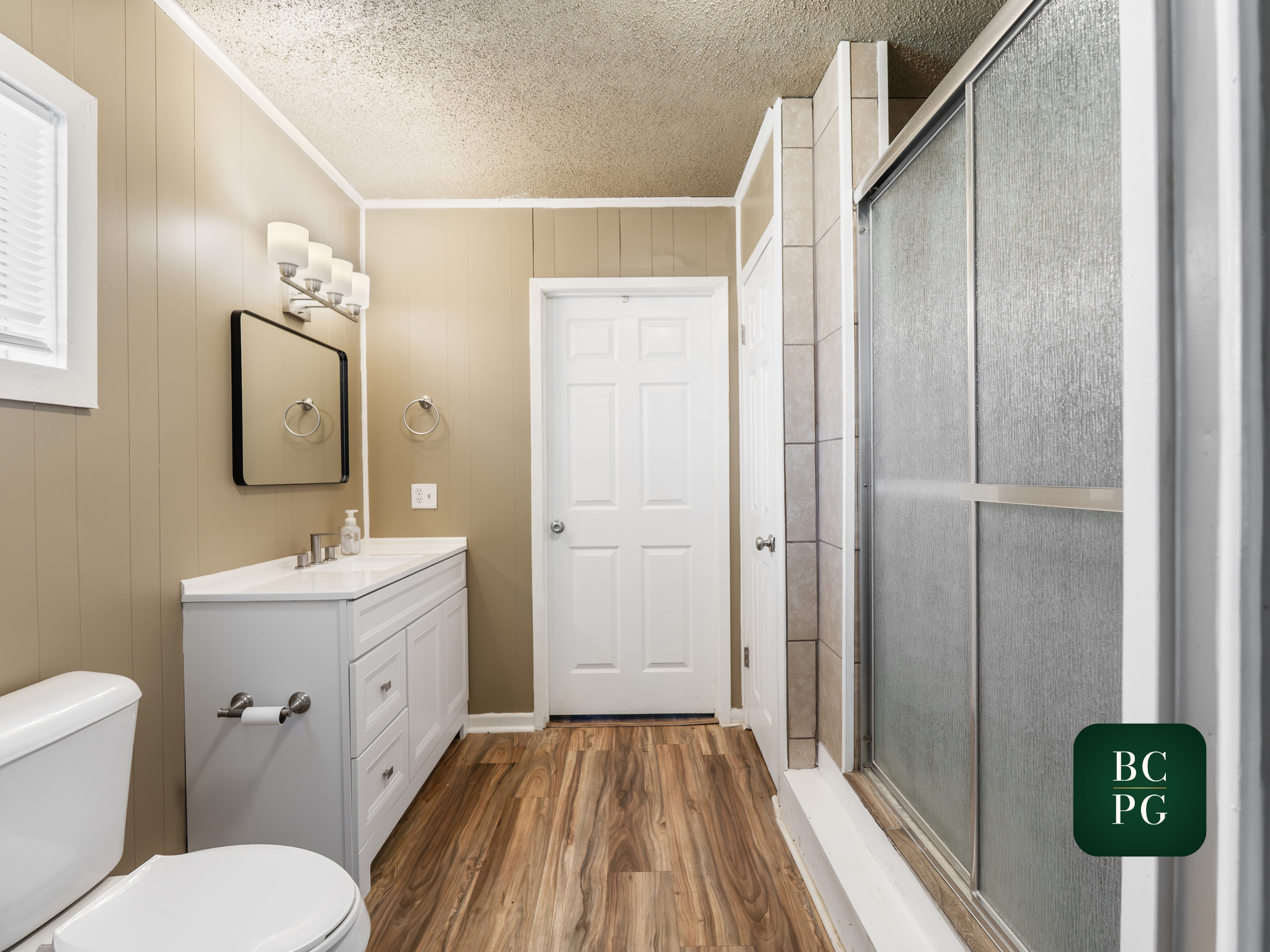 A small bathroom with beige paneled walls, a white vanity with a sink, a mirror, and wall-mounted light fixtures. There is a toilet in the foreground, a door, and a corner shower with frosted glass sliding doors. The floor is made of wood-look lamina