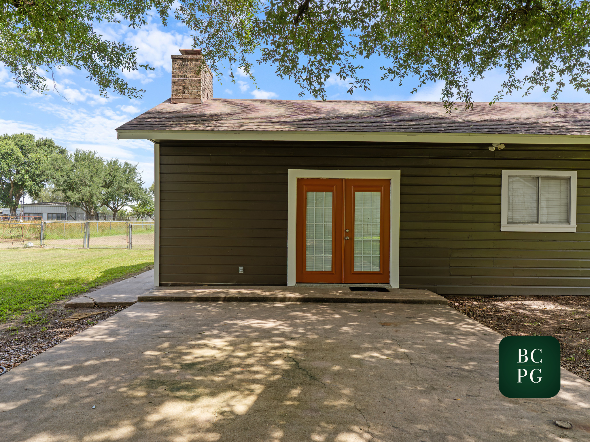A house with green siding, a red front door, and a brick chimney. There is a concrete porch and a yard with a chain-link fence, trees, and a blue sky with scattered clouds.