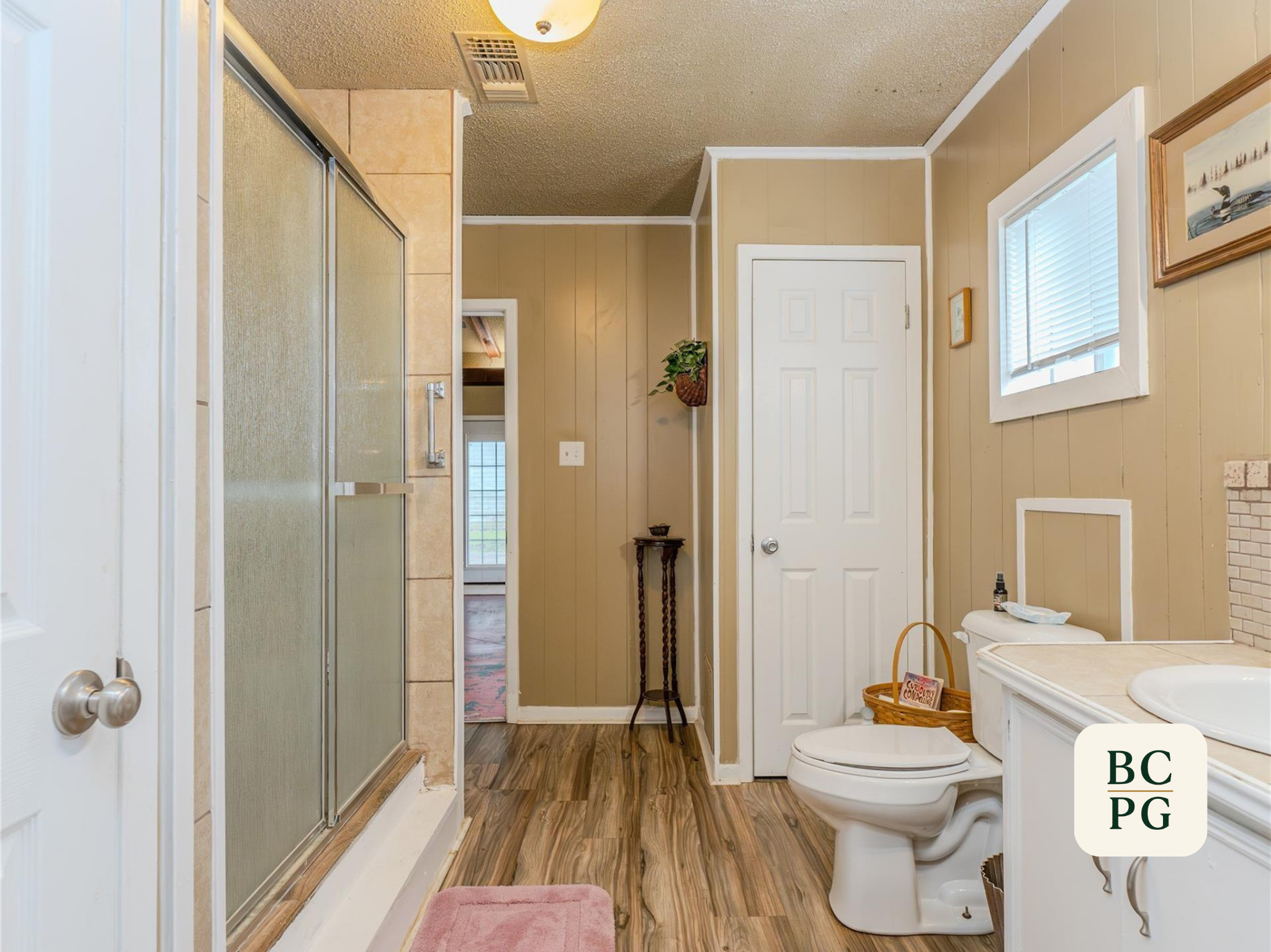 Bathroom with a shower, toilet, and sink, and decorated with pictures on the beige-paneled walls. There is a small pink bath mat on the wooden floor and a basket with magazines.
