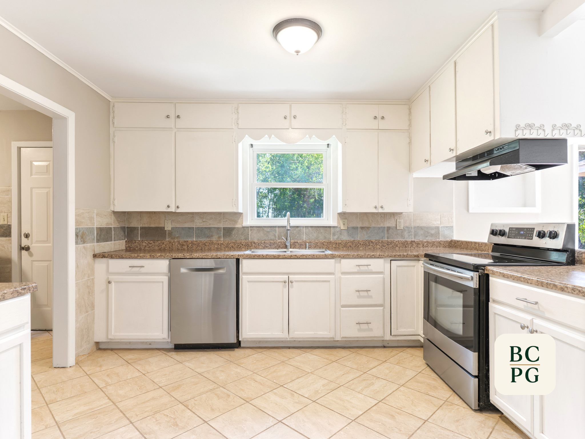 A bright kitchen with white cabinets, a window with a view of trees, a stove, and a beige tiled floor.