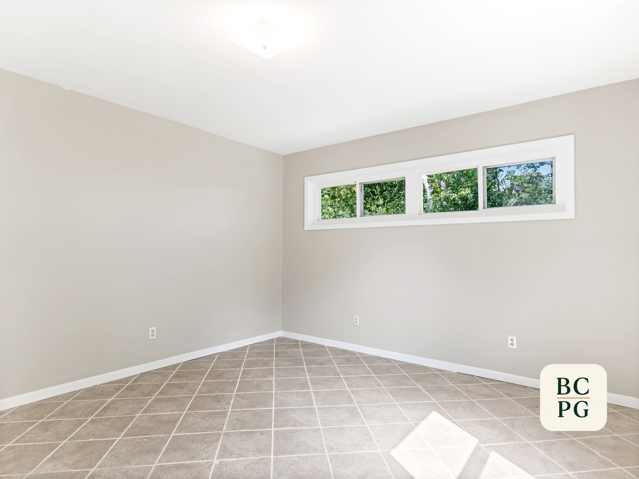 Empty room with beige walls, a row of narrow windows with greenery outside, and tiled floor.